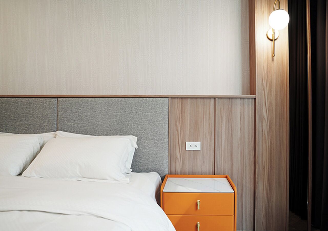 A hotel room with a bed, a nightstand with drawers, a wall-mounted light, and a power outlet.