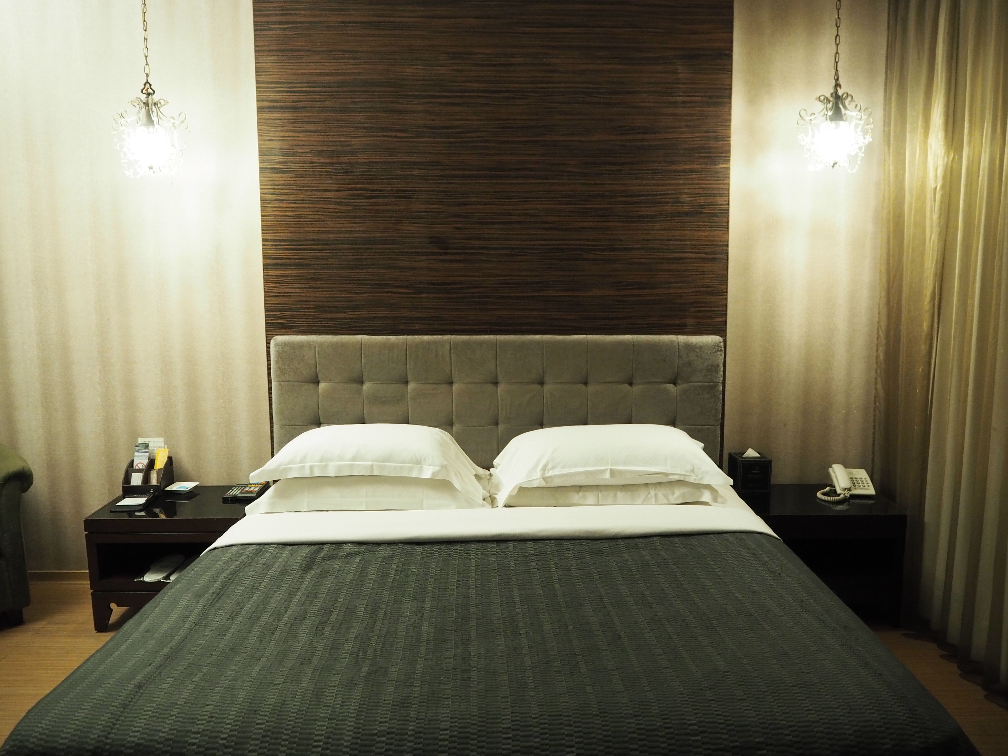 A neatly made bed with a grey headboard and white pillows, a bedside table with a phone and a lamp, and a wooden panel wall.