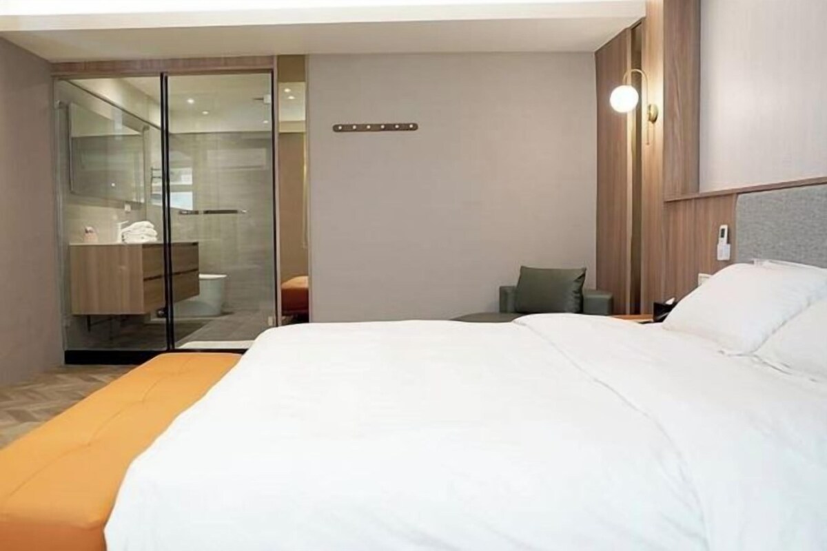 A modern hotel room with a large bed, a glass shower enclosure, a small seating area, and a wooden headboard.