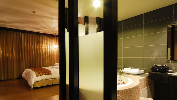 A hotel room with a bed, a bathroom with a sink and mirror, and a wooden floor. - The Riverside Hotel & Motel (Kaohsiung)