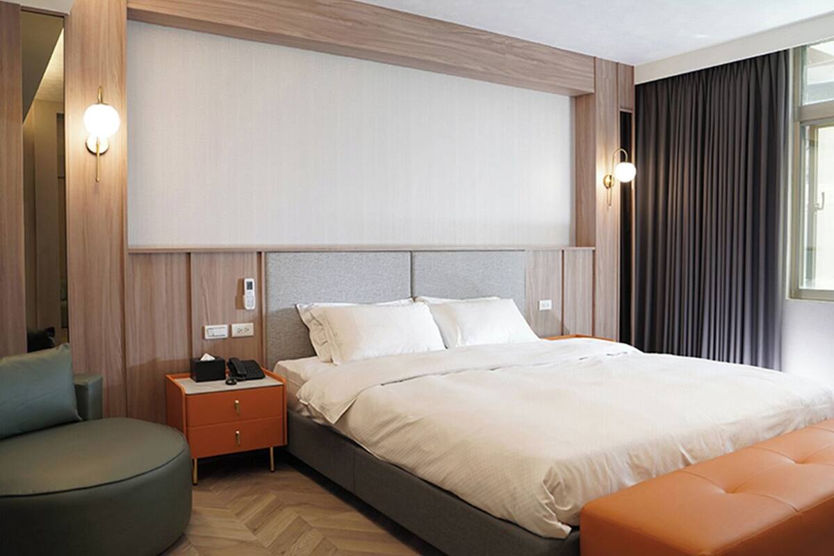 A modern hotel room with a large bed, a bedside table with a phone and a lamp, a round armchair, and a window with curtains.