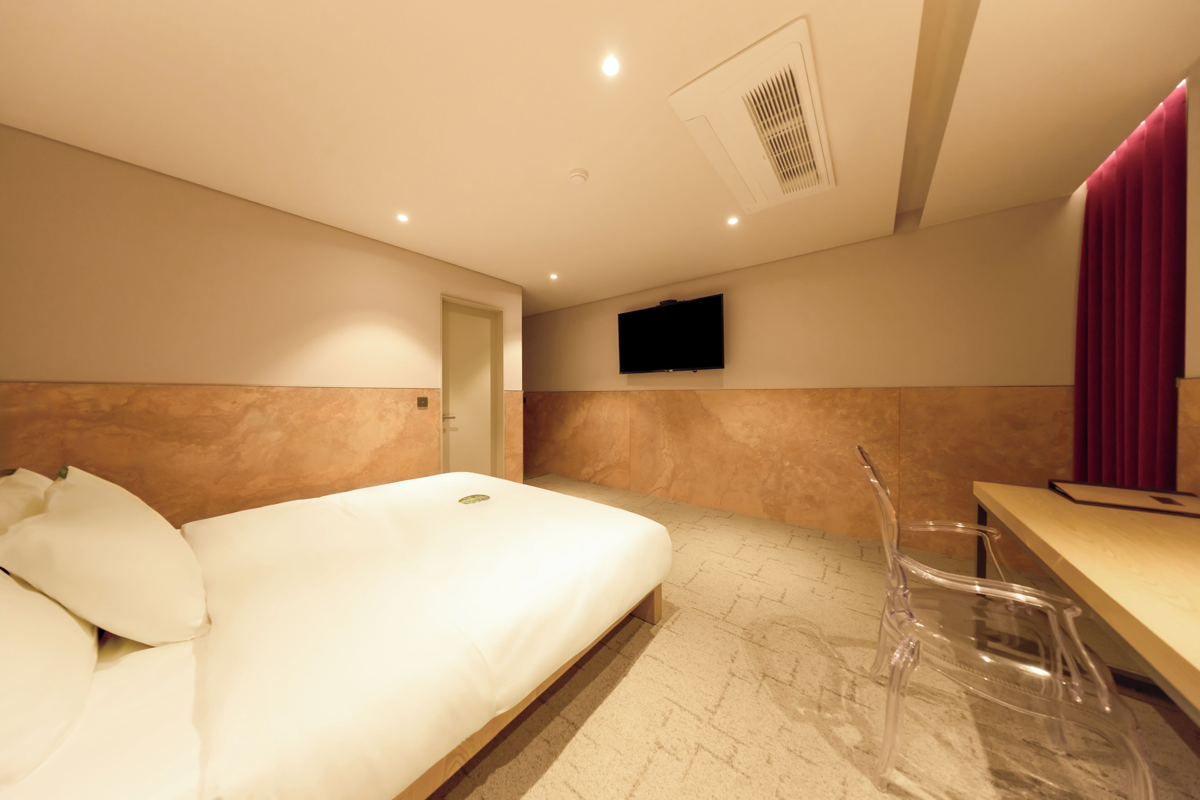 A hotel room with a bed, a desk, a chair, a TV mounted on the wall, and a door.