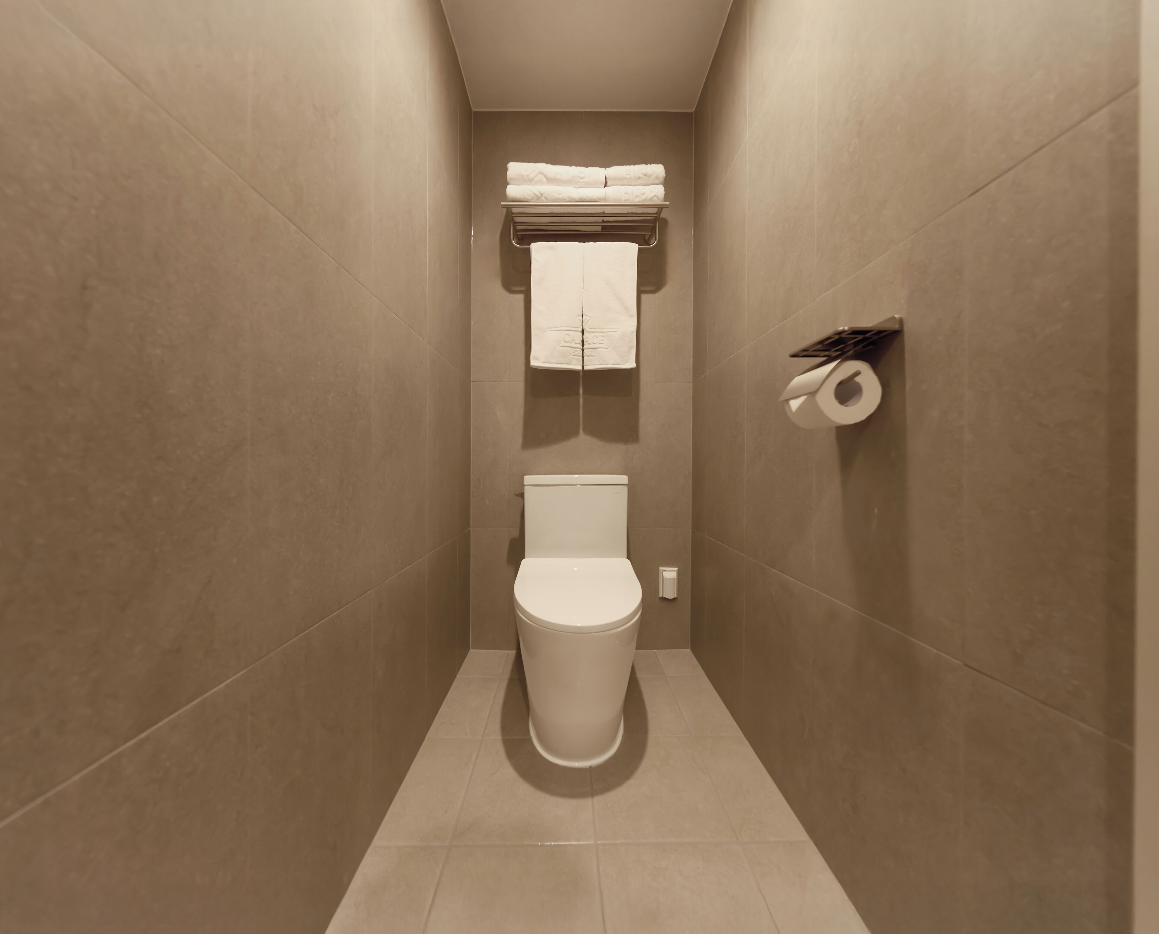A narrow, tiled bathroom with a toilet, a towel rack with folded towels, and a toilet paper holder.