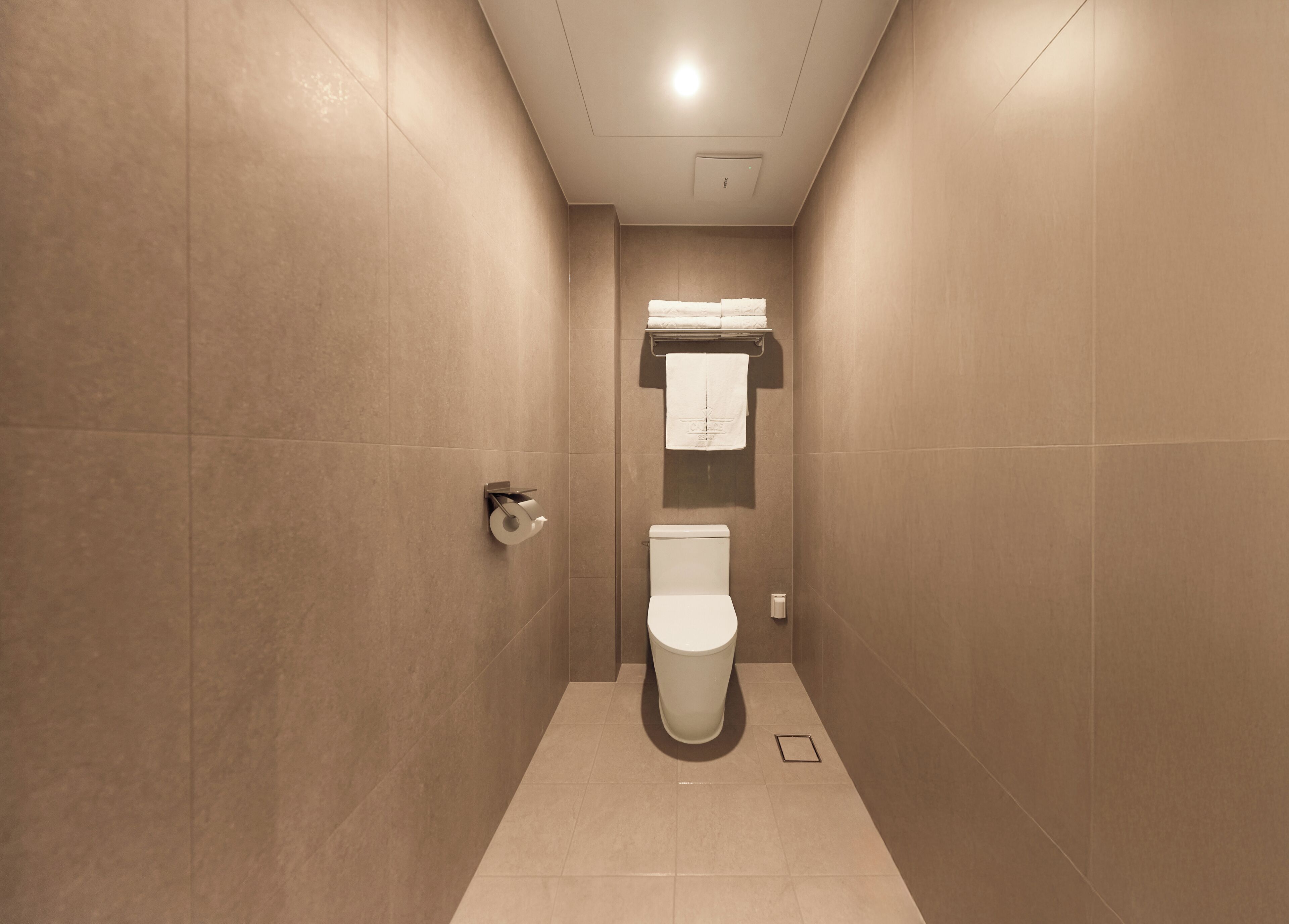 A modern bathroom with beige tiles, a toilet, a towel rack, and a paper towel dispenser.