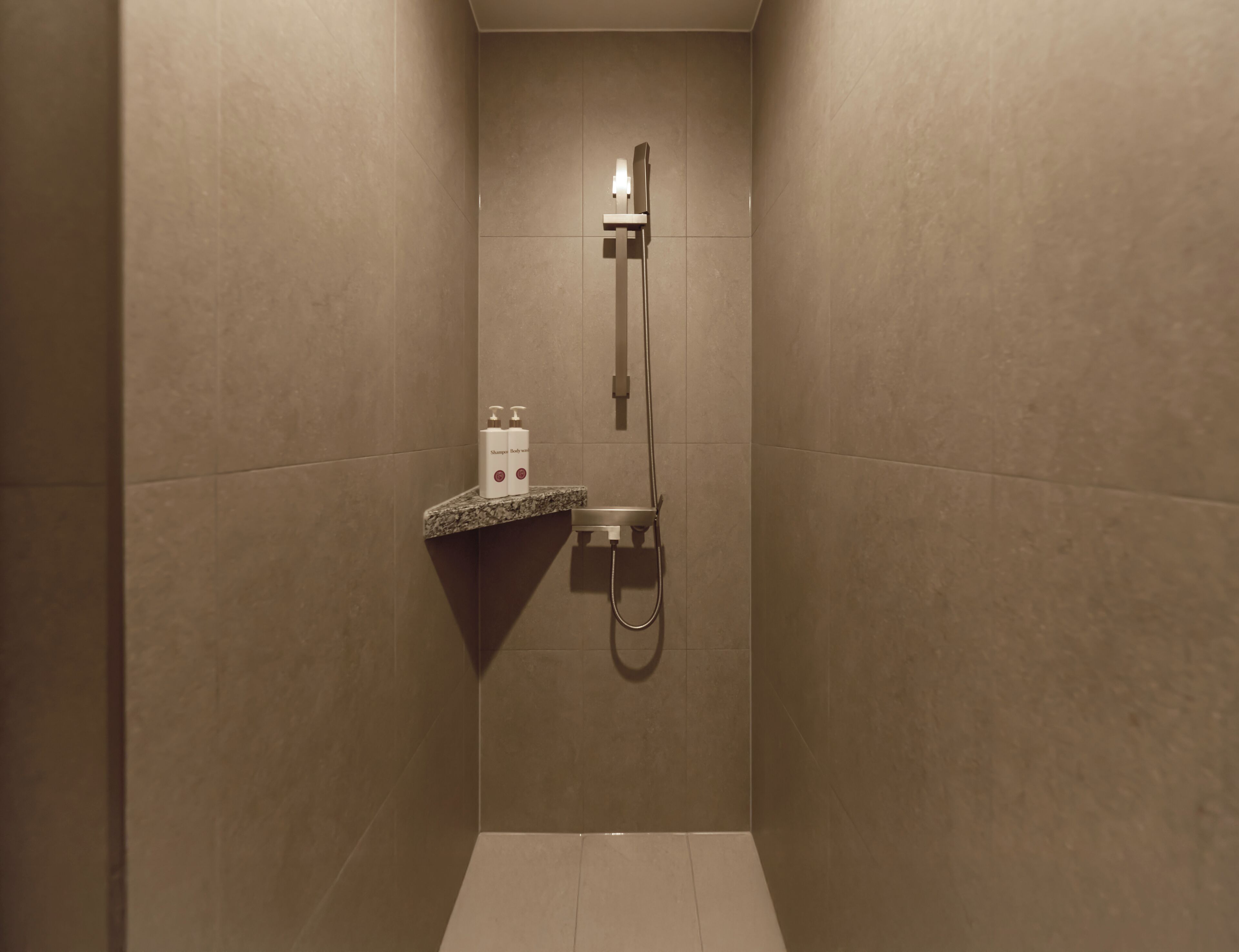 Ein kleiner, abgeschlossener Duschbereich mit Handbrause und am Wand montiertem Wasserhahn. Es gibt ein Regal mit zwei Shampoo- und Conditionerflaschen.