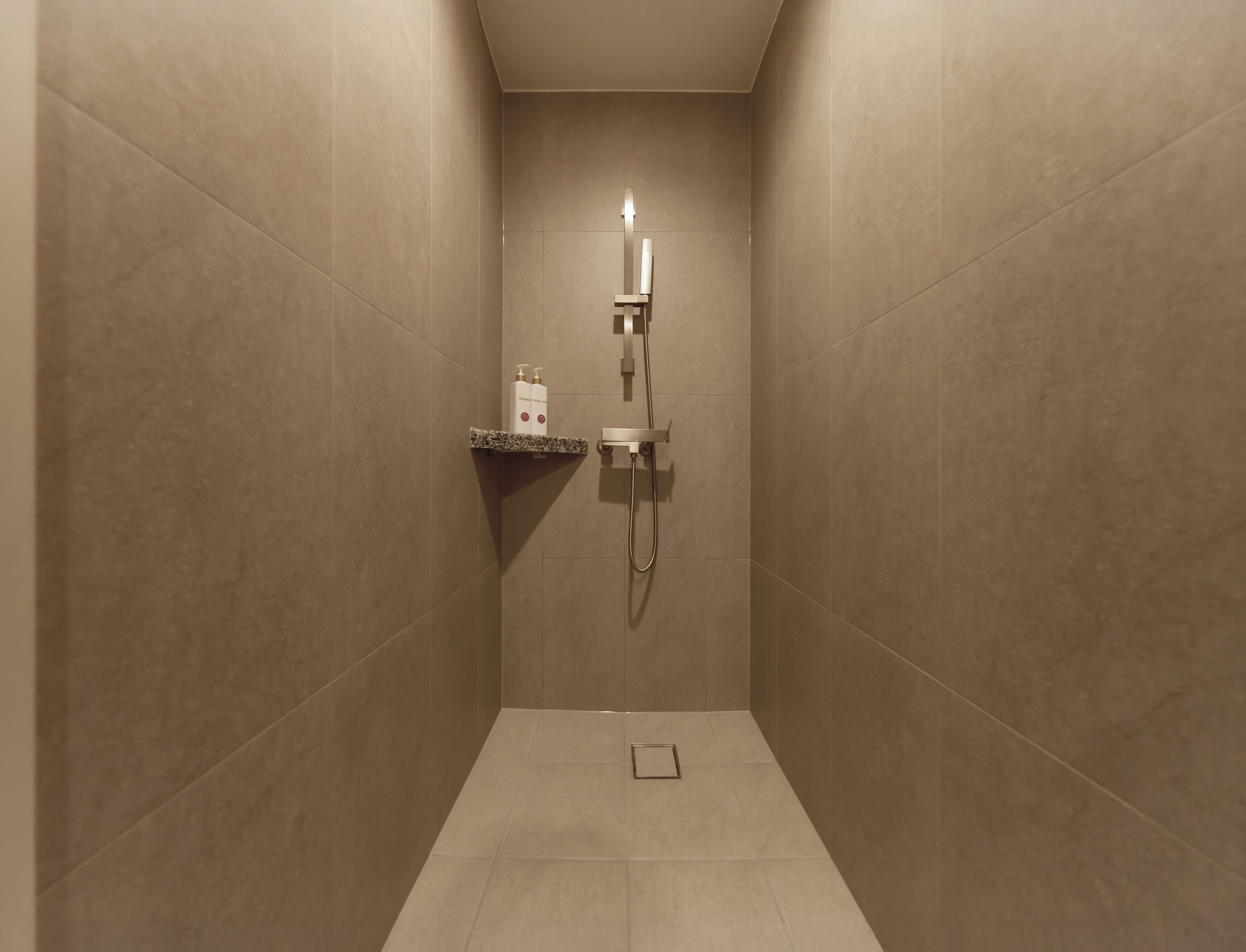 A narrow shower area with beige tiles, a built-in shelf with toiletries, and a handheld showerhead.