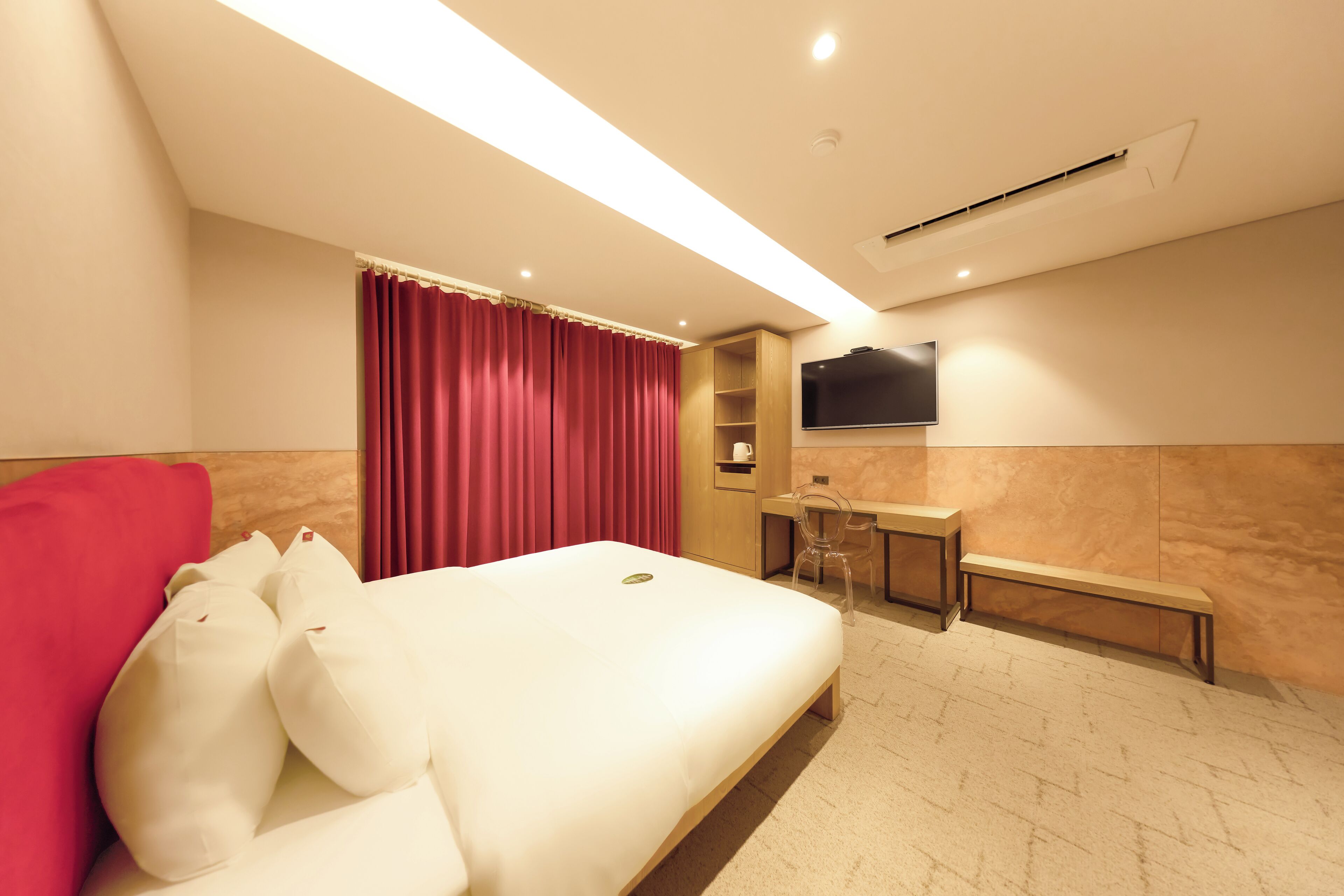 A hotel room with a bed, a desk, a chair, a TV, and a bench.