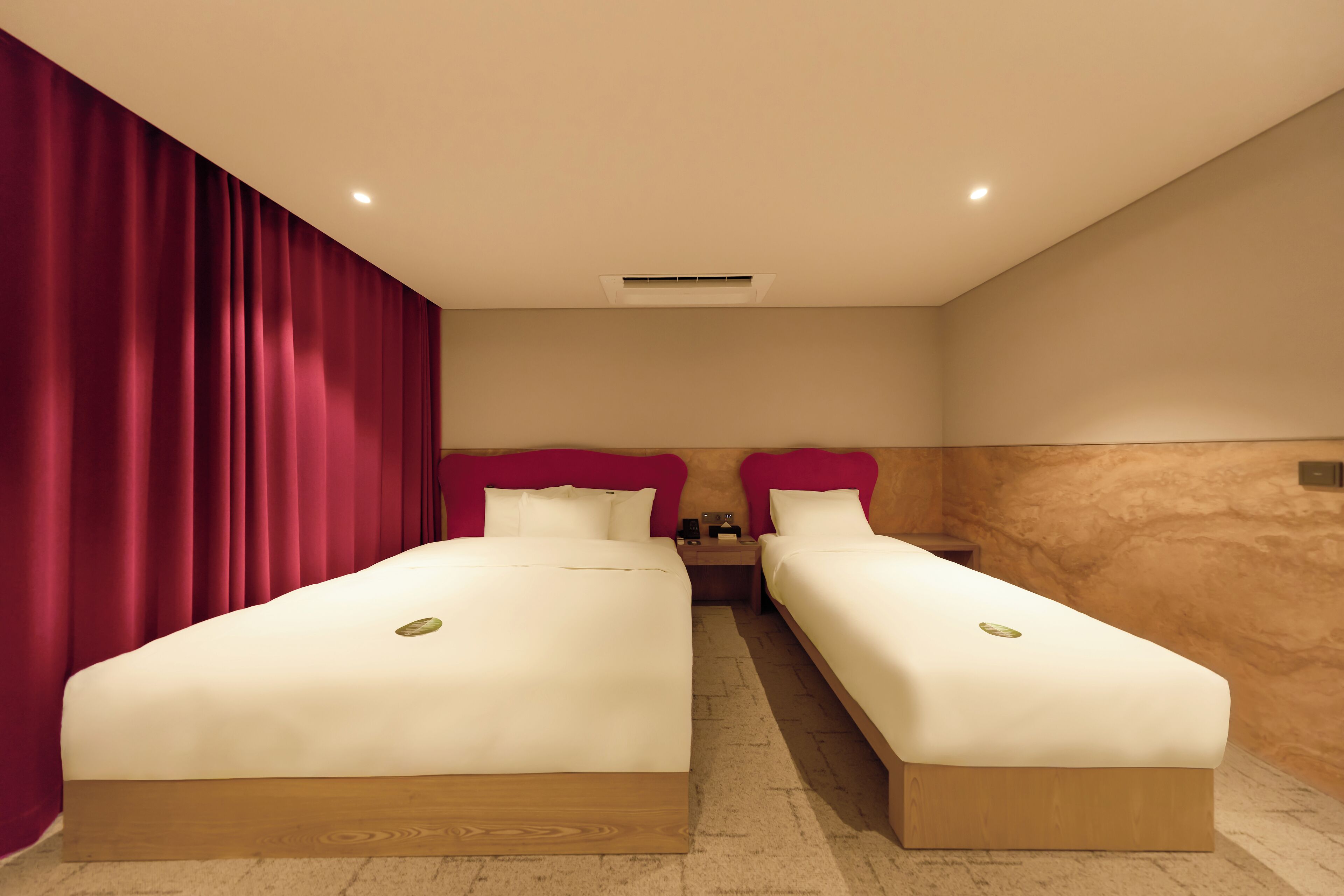 A hotel room with two beds, red curtains, beige walls, and a wooden floor.