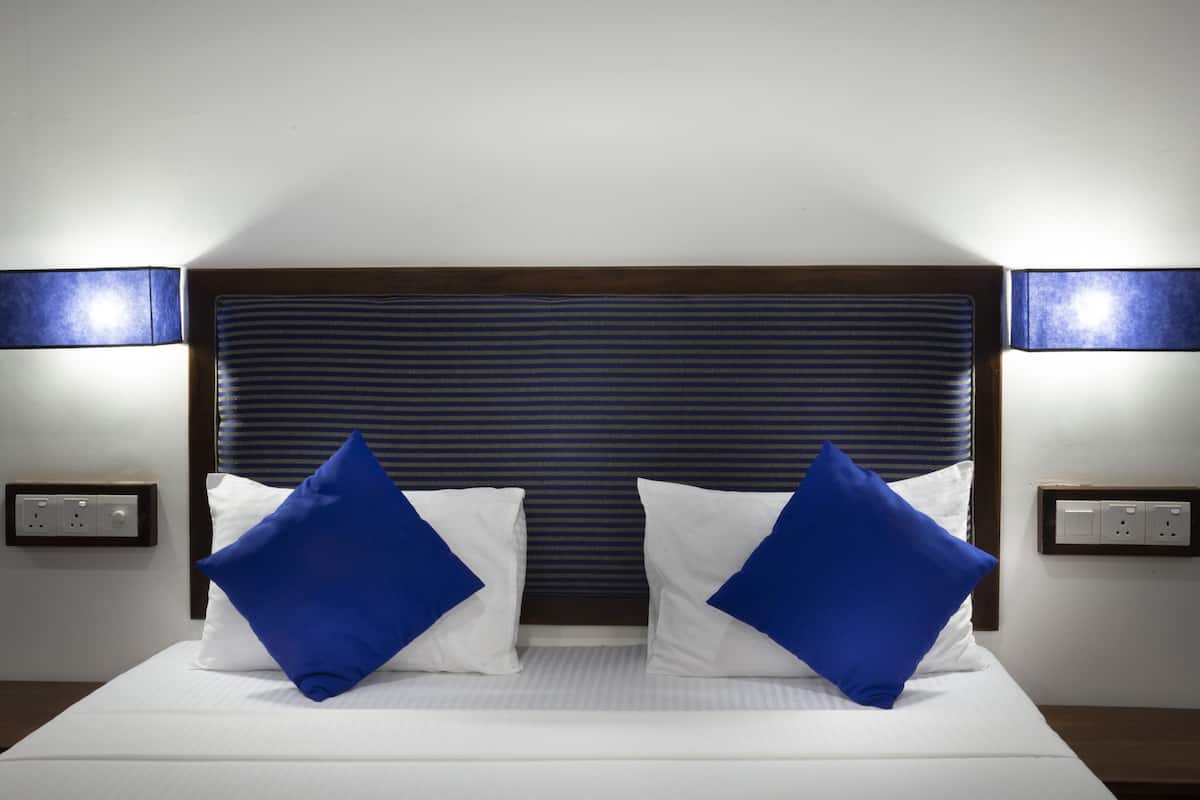 Una cama bien hecha con sábanas blancas y dos almohadas azules, un cabecero oscuro y dos lámparas fijadas a la pared.