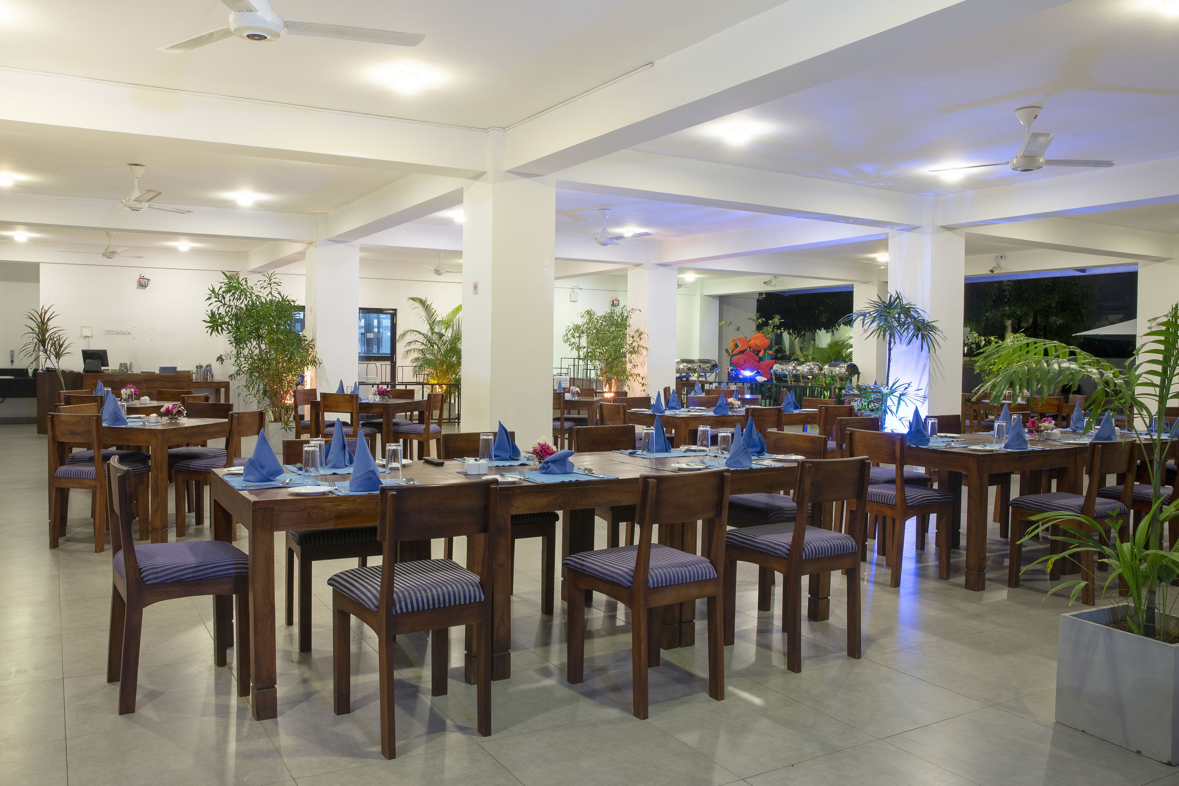 A spacious dining area with multiple tables and chairs, ceiling fans, and potted plants.