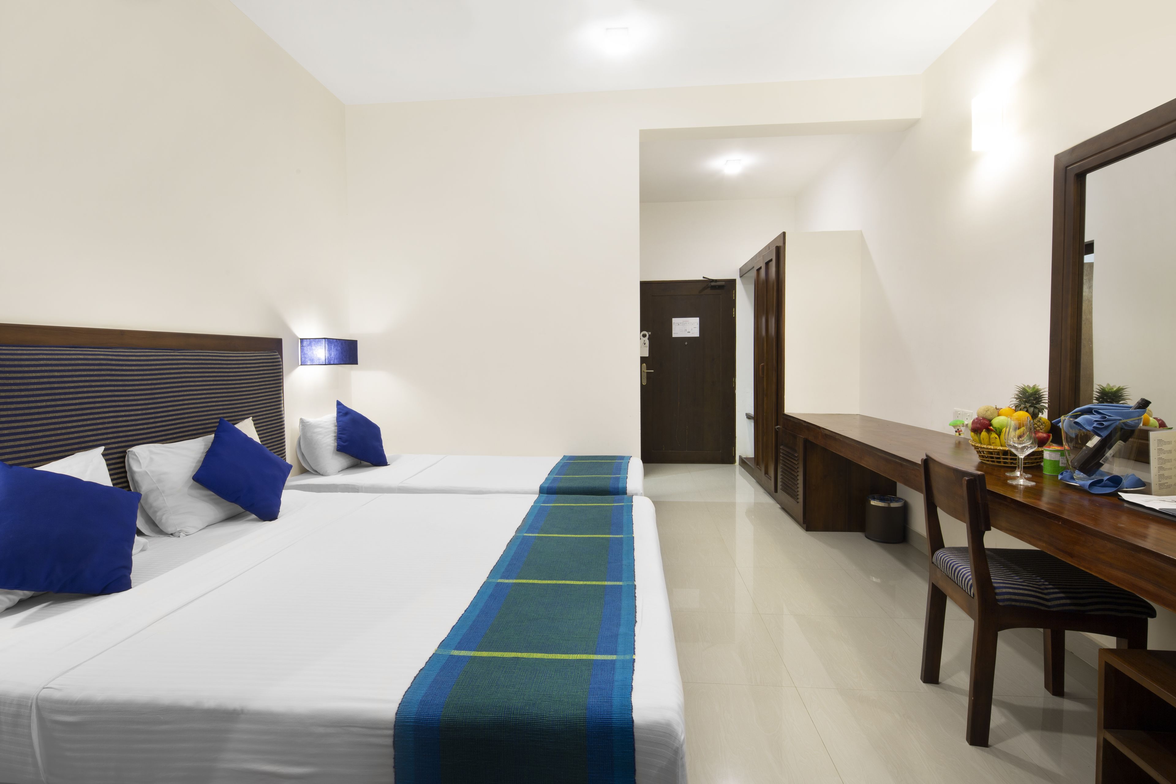 A hotel room with two beds, a desk, a chair, a mirror, and a small table with a fruit basket.