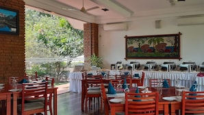 A restaurant with multiple tables set for dining, a large painting on the wall, and a view of greenery outside.