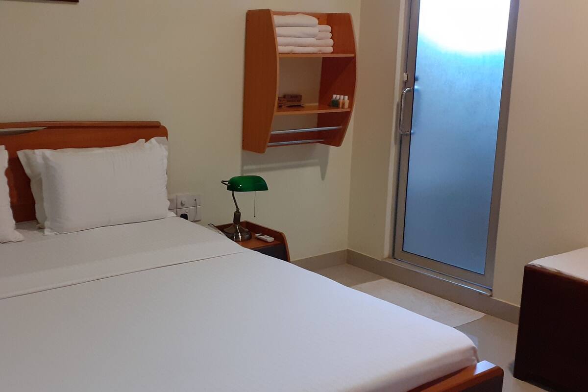 Habitación de hotel con cabecera de madera, ropa de cama blanca, mesita de noche con lámpara y ventana con vistas.