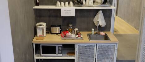 A kitchenette with a coffee maker, toaster, microwave, and sink. There are shelves above holding dishes and a sign indicating "Easy Breakfast Self Service 07:00-10:00".
