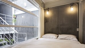 A bedroom with a large window, a bed with white bedding, and a concrete wall with a light fixture. - On the bed Hostel (Bangkok)