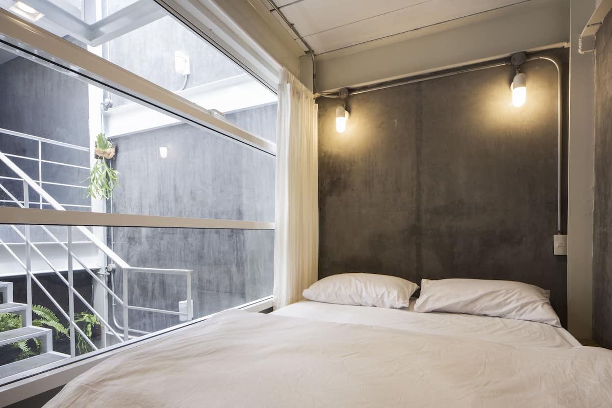 A bedroom with a large window, a bed with white bedding, and a concrete wall with a light fixture.