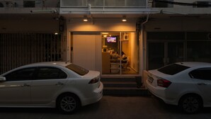 A modern building with a unique facade featuring vertical panels. There is a visible entrance with a door and a small window, and a car parked in front. - On the bed Hostel (Bangkok)