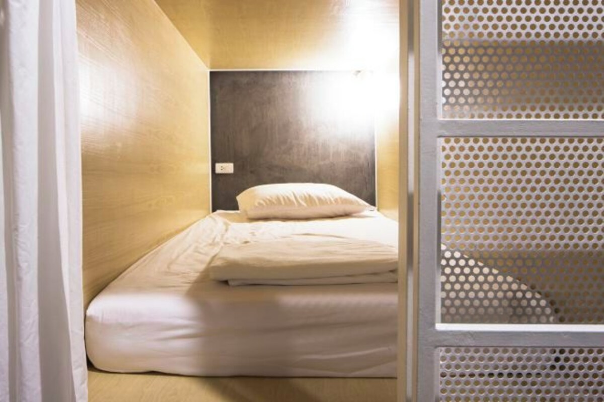 Una habitación pequeña y estrecha con una cama individual, cabecera y ventana con mosquitera.