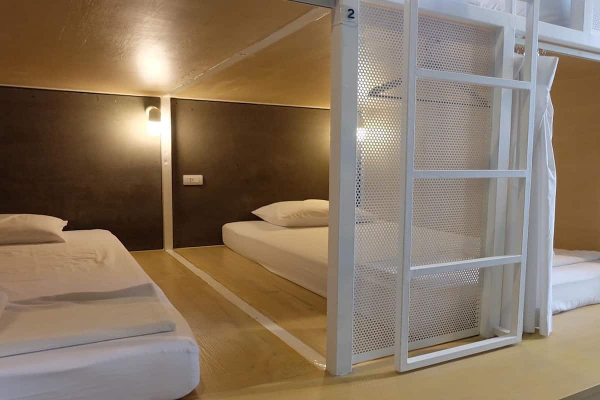 A hotel room with two beds, a wooden floor, a wall-mounted light, and a white partition with a mesh design.