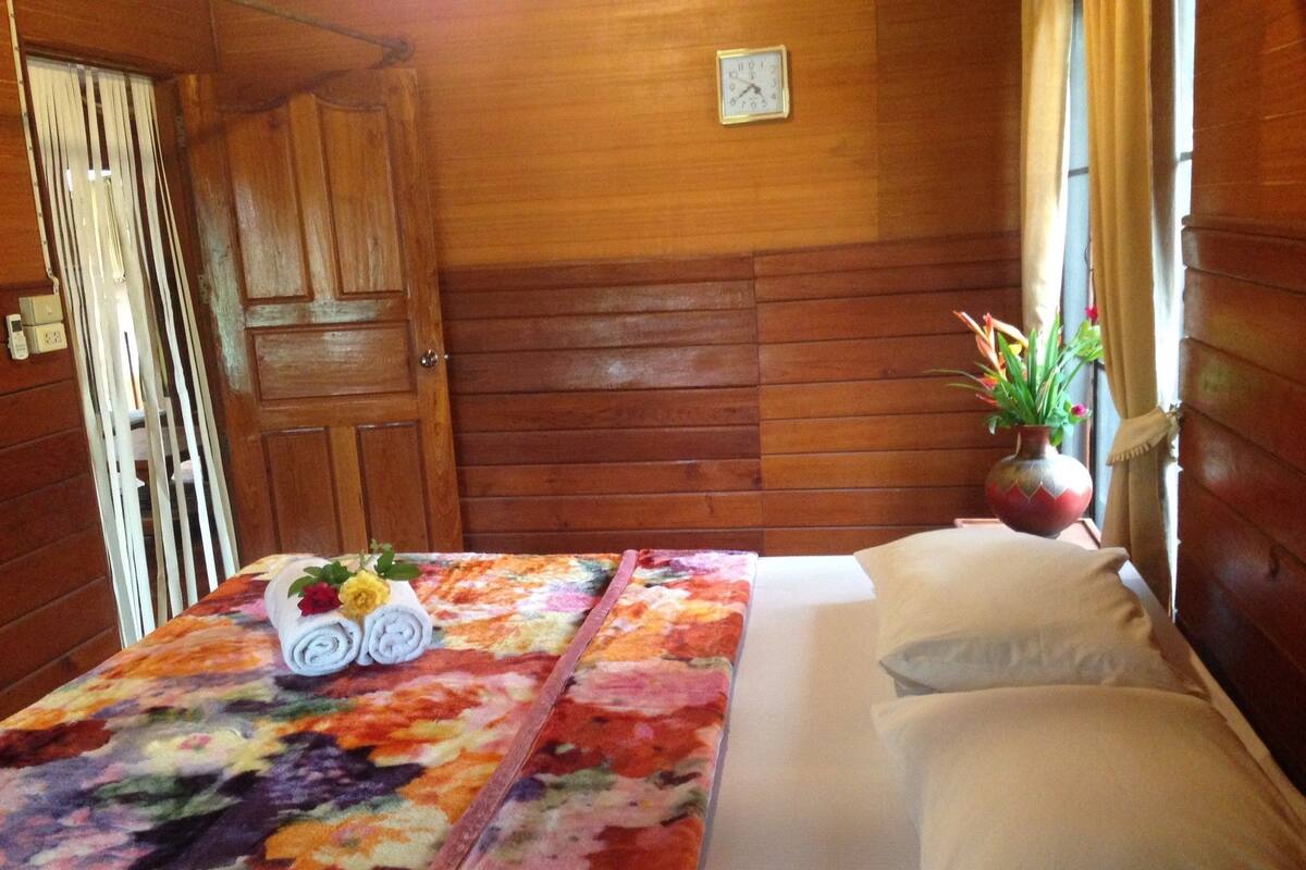 Una habitación con paneles de madera, una cama con sábanas blancas, un mantel con estampado floral y un reloj en la pared.