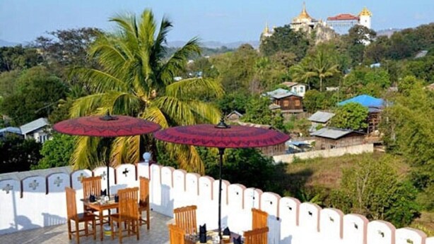 A terrace with wooden tables and chairs, red umbrellas, and a view of a temple and houses on a hill. - Hotel Myat Nan Taw (Loikaw)