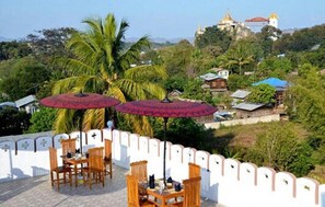 Una terraza con mesas y sillas de madera, sombrillas rojas y vistas a un templo y casas en una colina.