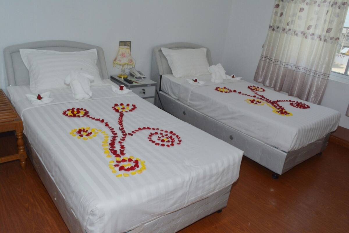 Ein Hotelzimmer mit zwei Betten, jeweils mit weißer Bettwäsche und floralen Verzierungen. Zwischen den Betten befindet sich ein Nachttisch mit einer Lampe und einem Telefon.