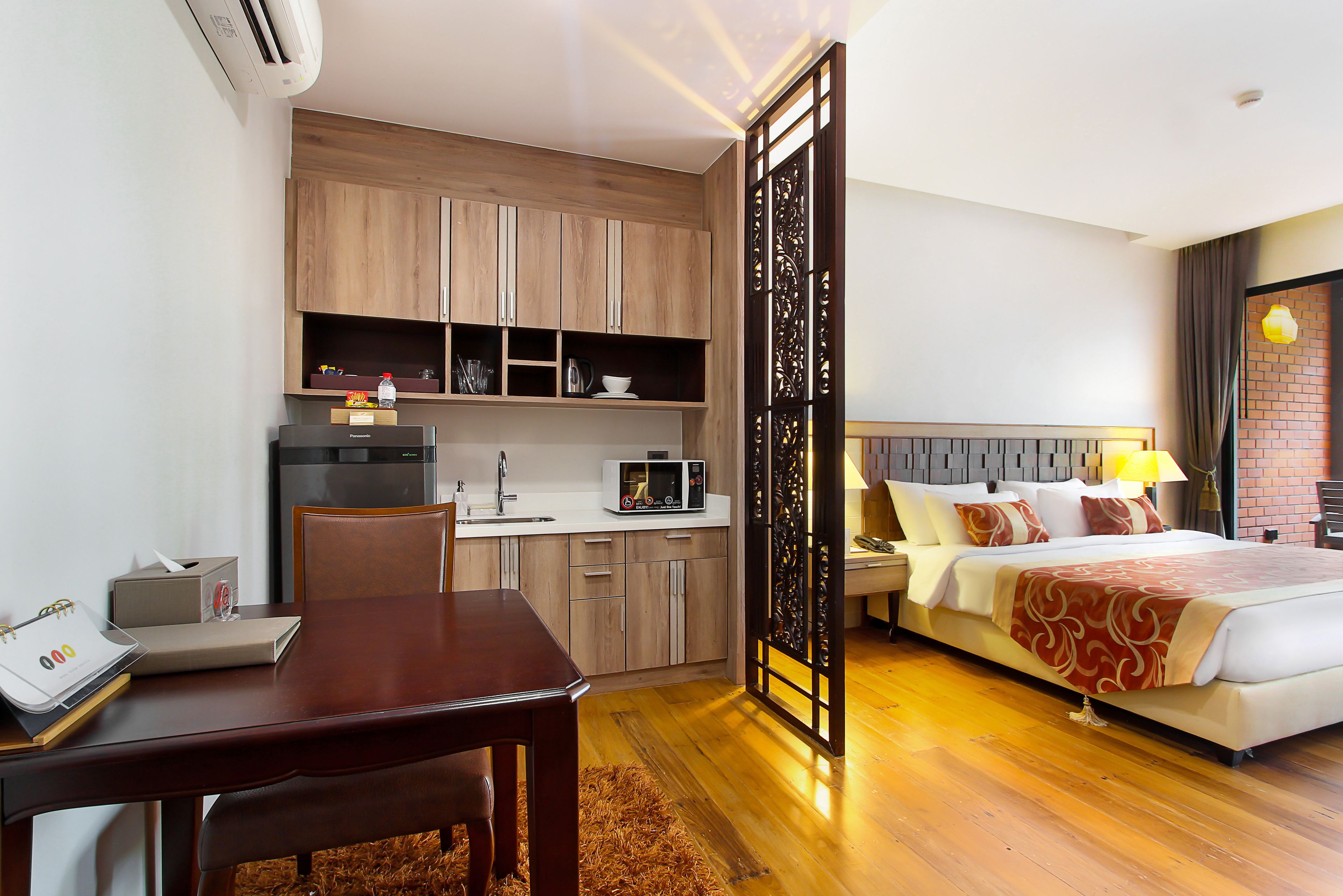 Habitación de hotel con escritorio de madera, kitchenette y cama con colcha roja y blanca.