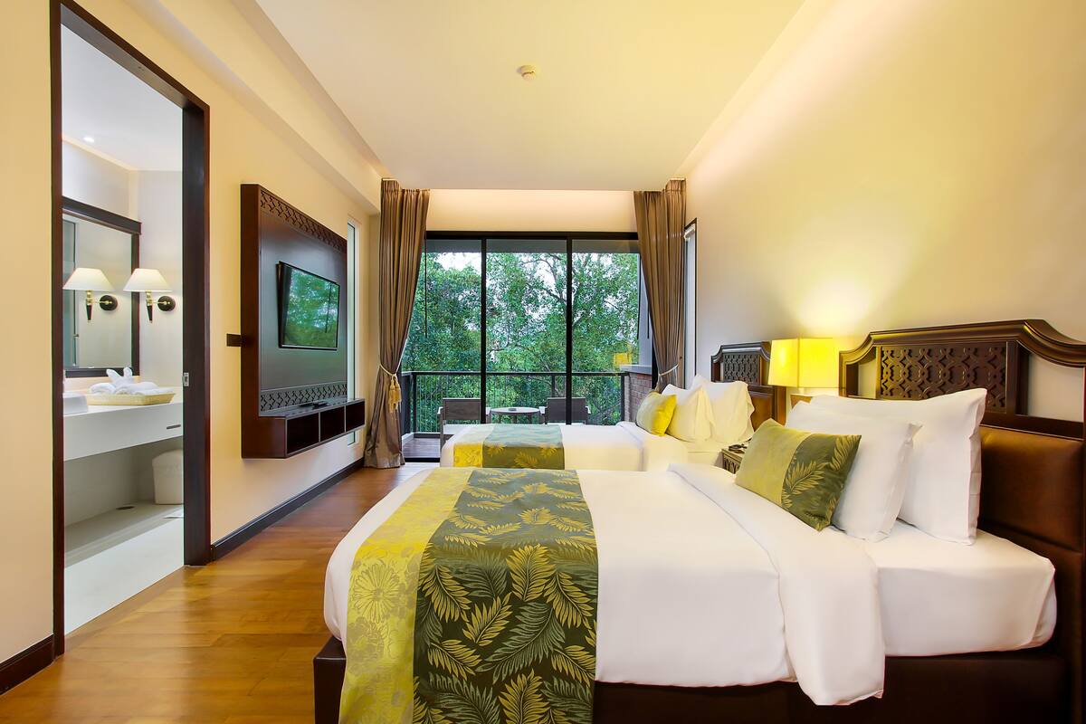 Habitación de hotel con una cama grande, televisión, balcón con vistas a árboles y baño con espejo y toallero.