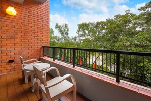 A balcony with a brick wall, a black railing, and a view of trees. There are two wooden chairs and a small table.