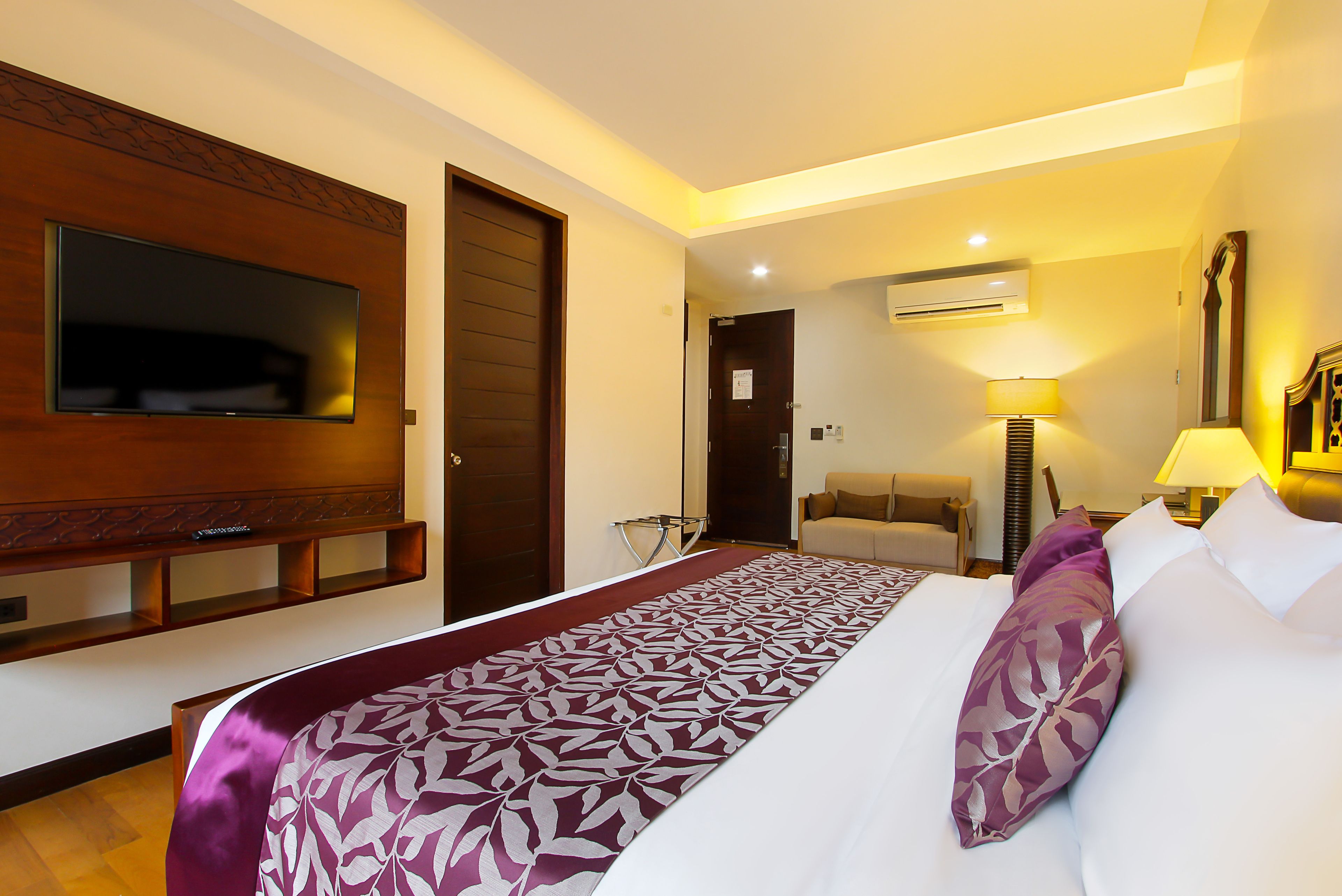 A hotel room with a large bed, a television mounted on the wall, a sofa, and an air conditioning unit.