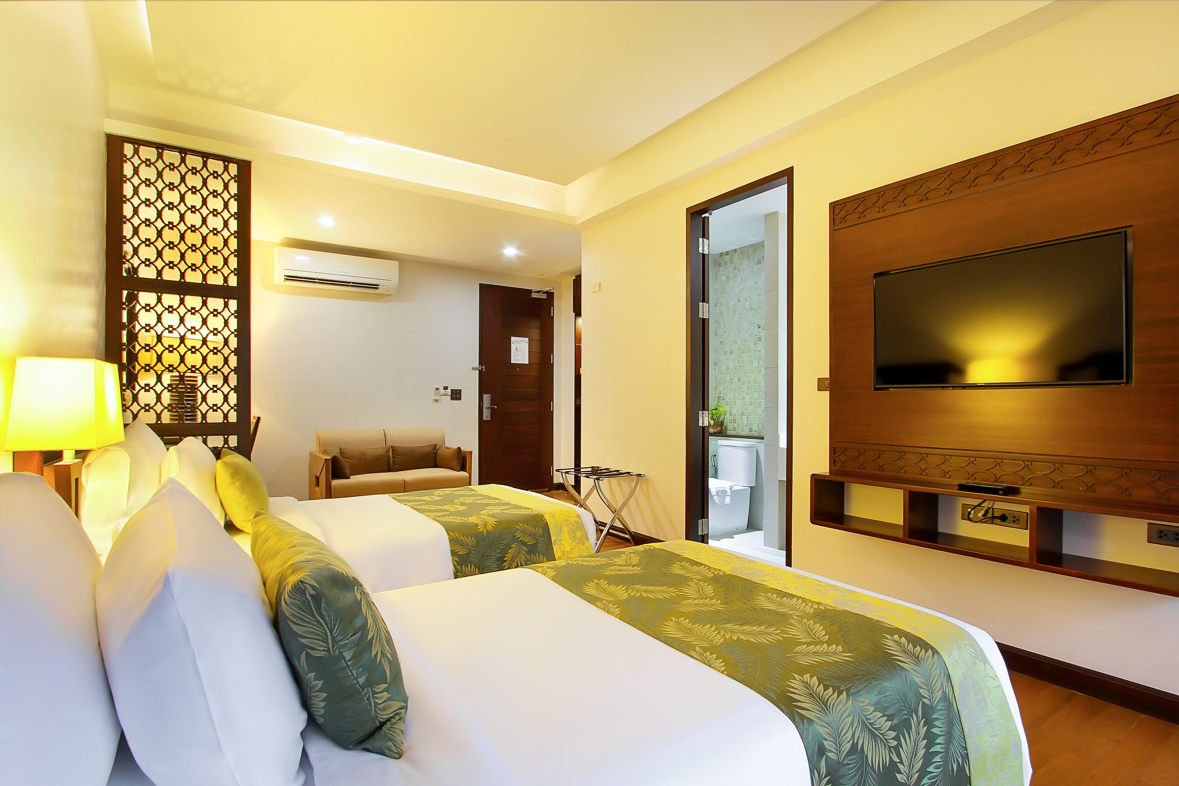 A hotel room with two beds, a TV, an air conditioning unit, and a small seating area.