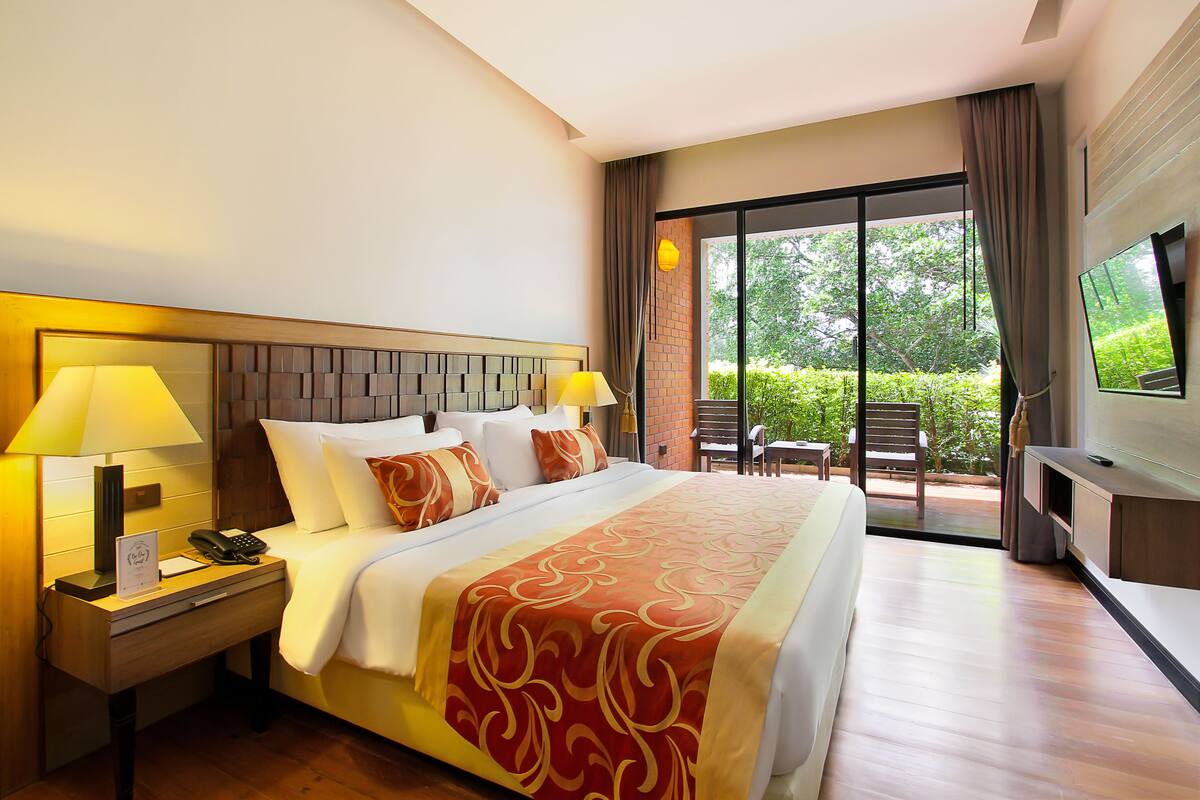 Habitación de hotel con una cama grande, dos lámparas de noche, un televisor y un balcón con vistas a la vegetación.