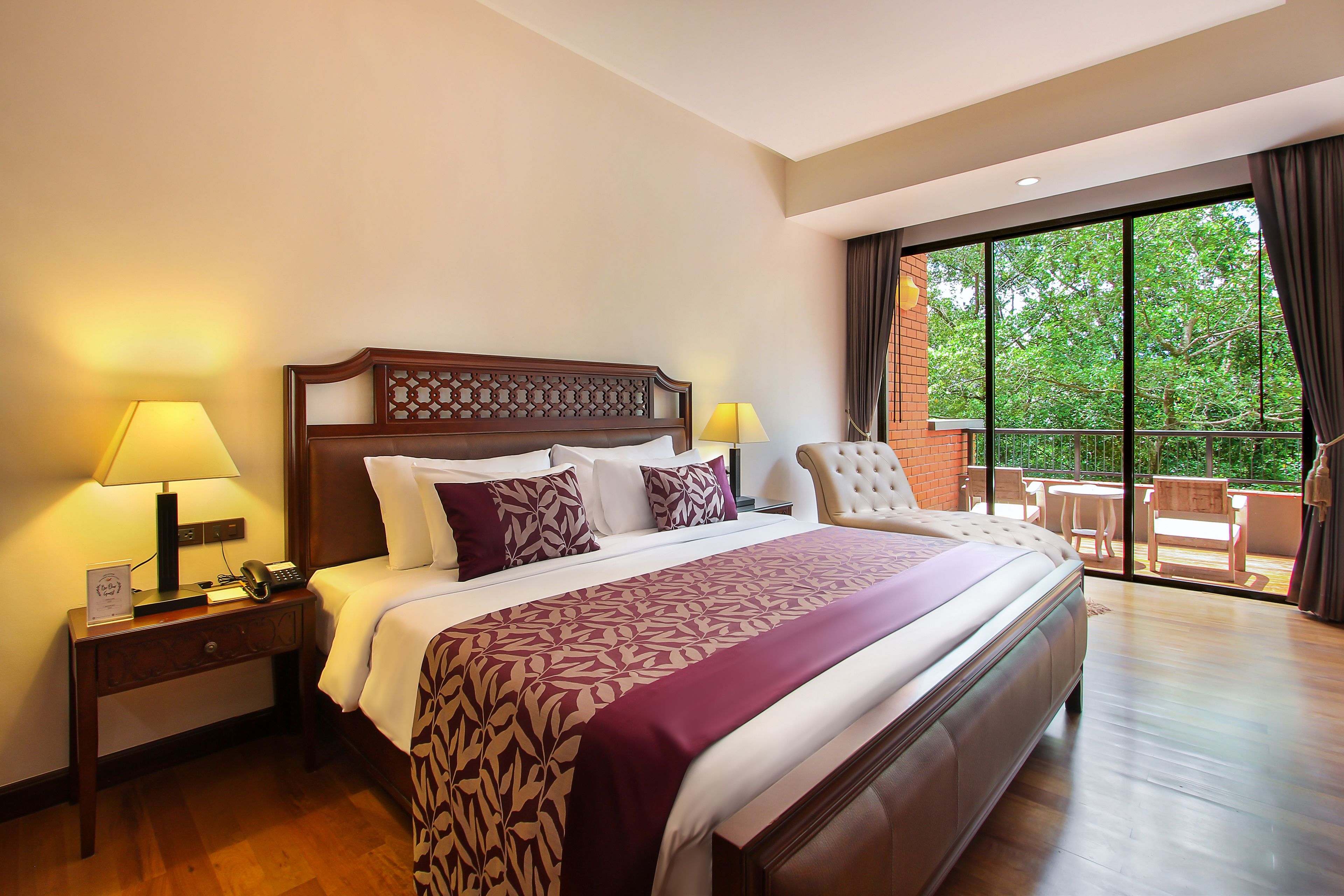 A hotel room with a large bed, two bedside tables with lamps, a view of the outdoors through a large window, and a balcony with a table and chairs.