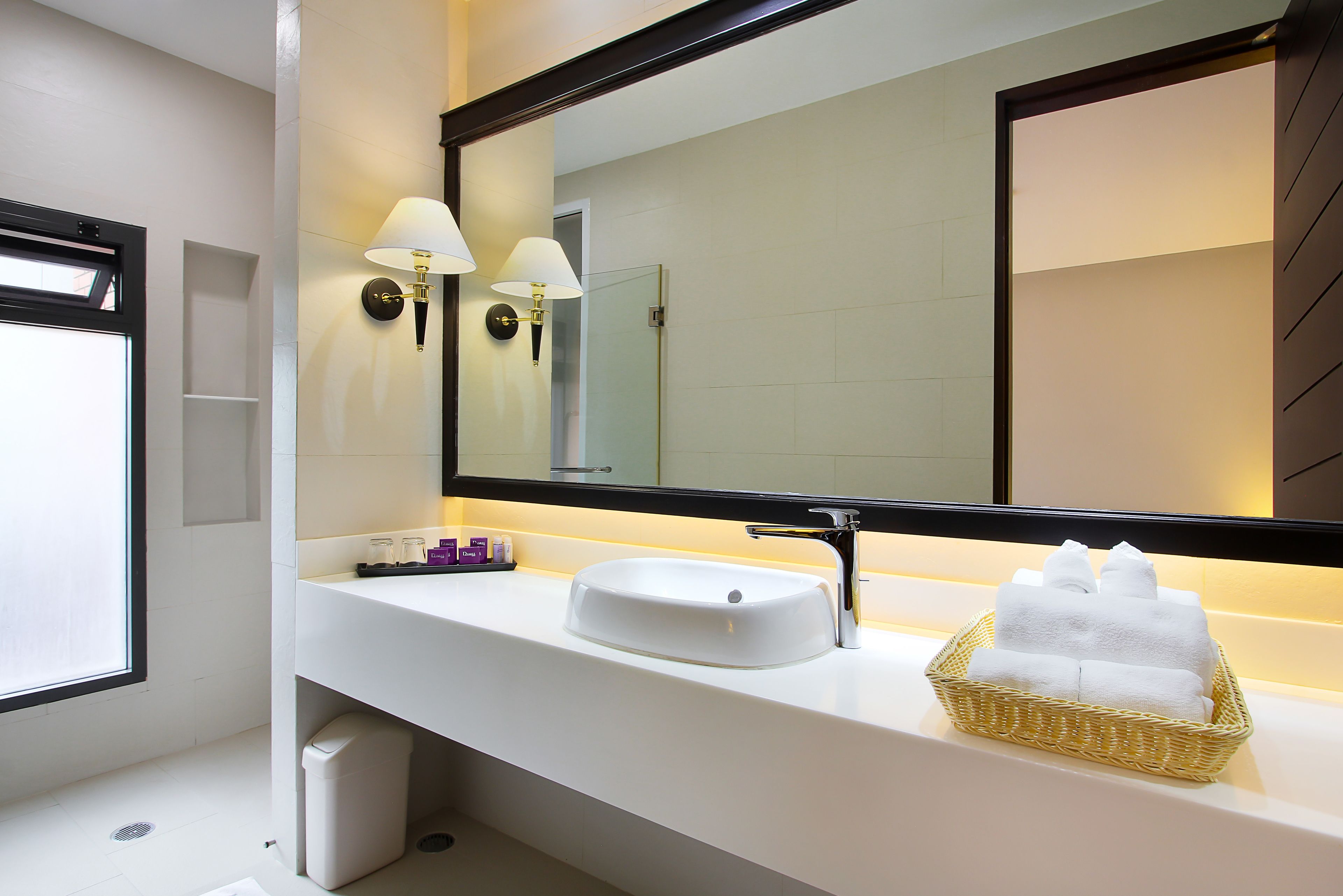 A modern bathroom with a white sink, two wall-mounted lamps, a large mirror, and a basket of towels.