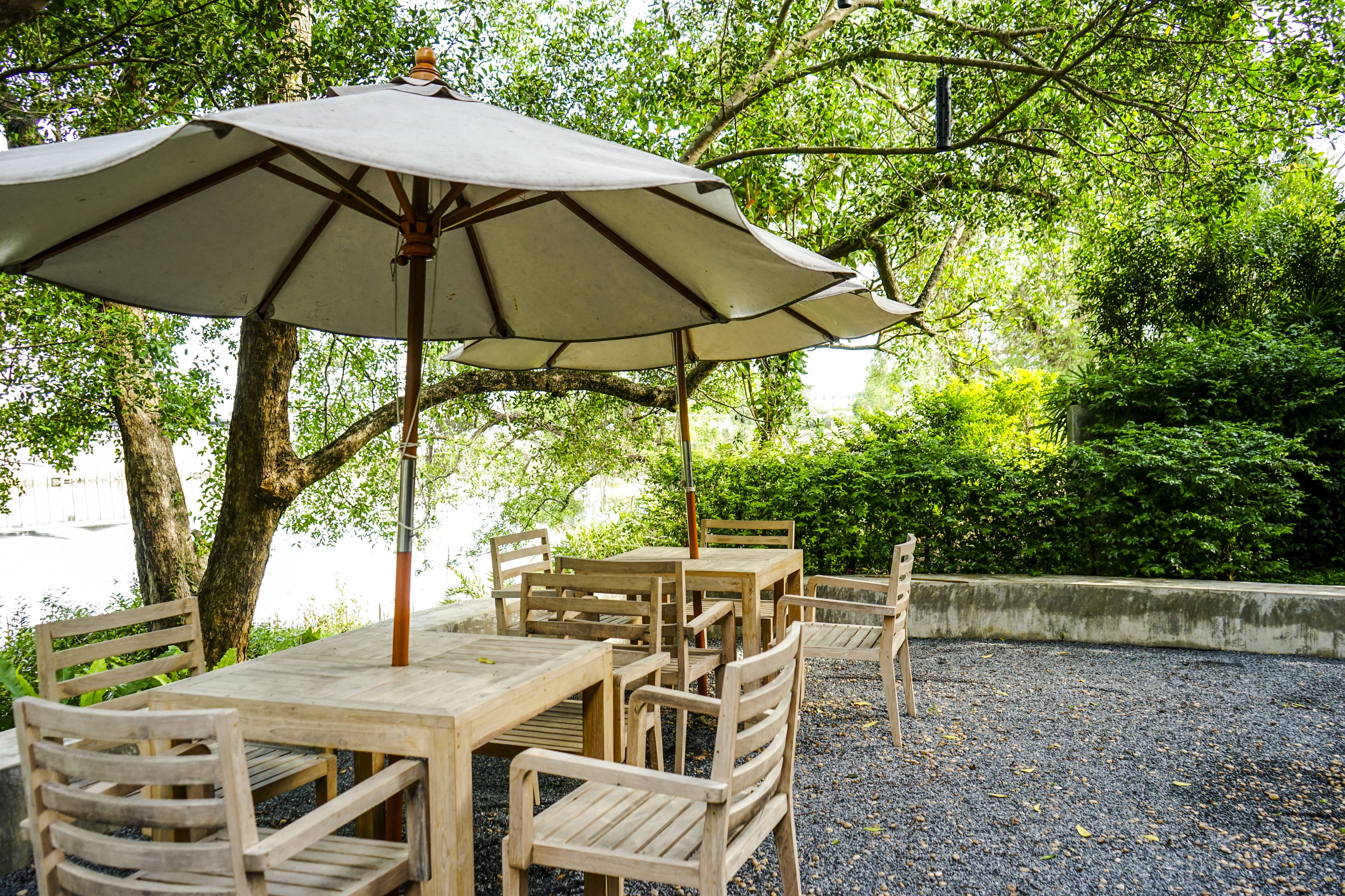 An outdoor dining area with wooden tables and chairs, a large umbrella, and a gravel ground.