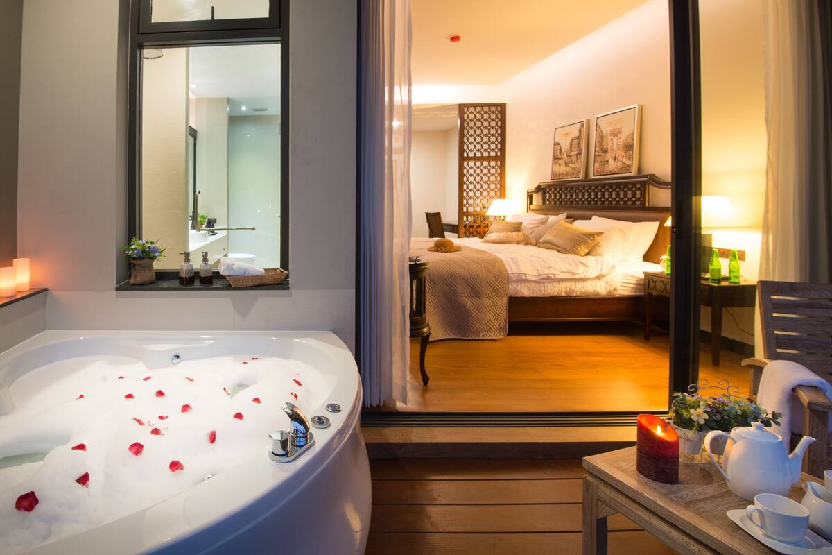 Una habitación de hotel con una bañera grande llena de burbujas y pétalos de rosa, una cama con cabecera de madera, una mesita de noche con tetera y tazas, y un espejo en la pared.