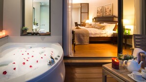 Una habitación de hotel con una bañera grande llena de burbujas y pétalos de rosa, una cama con cabecera de madera, una mesita de noche con tetera y tazas, y un espejo en la pared.