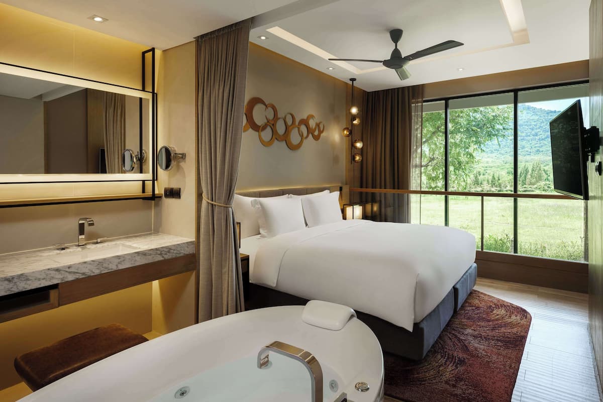 A modern hotel room with a large bed, a separate bathroom with a bathtub, a large mirror, and a view of a green landscape through the window.