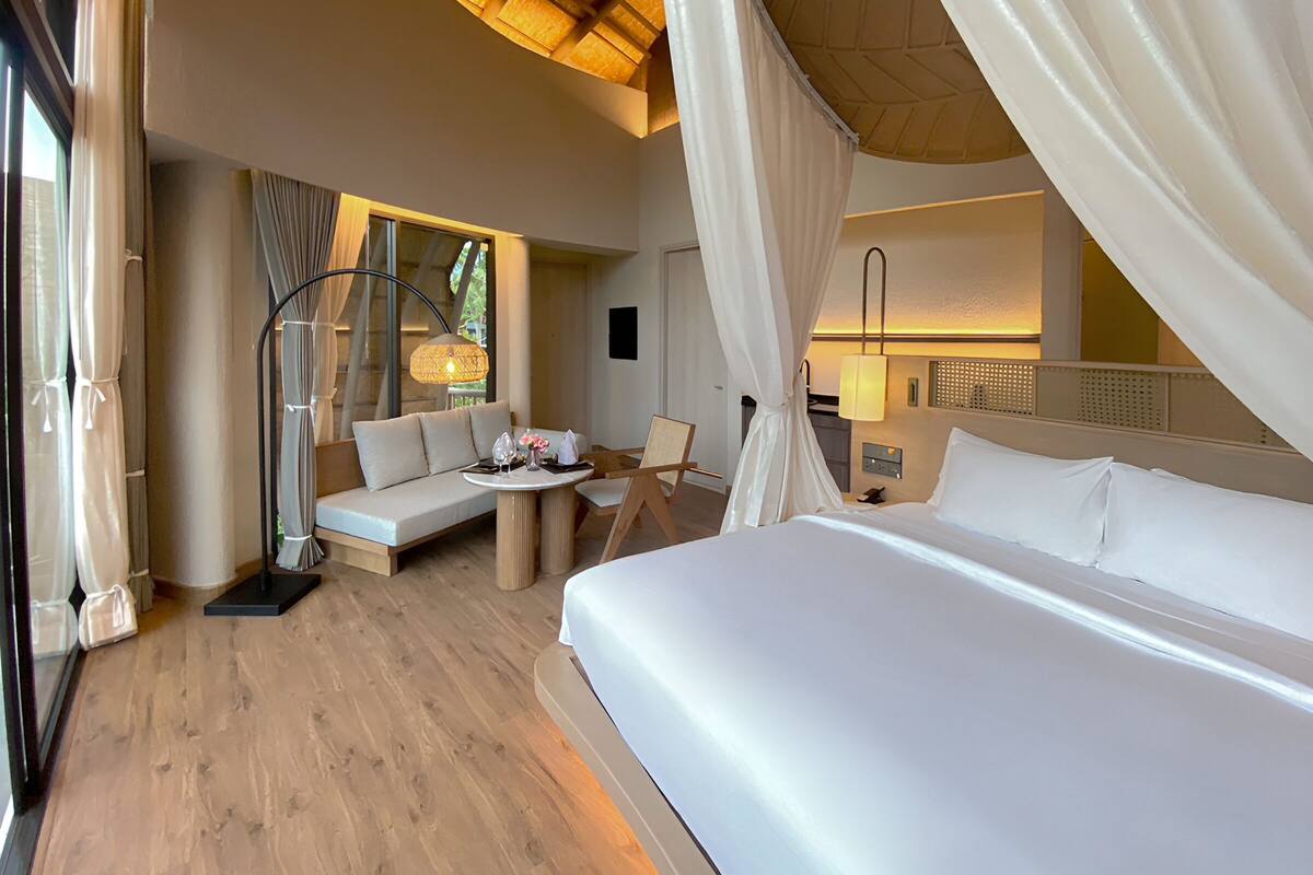 A bedroom with a large bed, a sofa, a small table with a lamp, and a window with curtains.