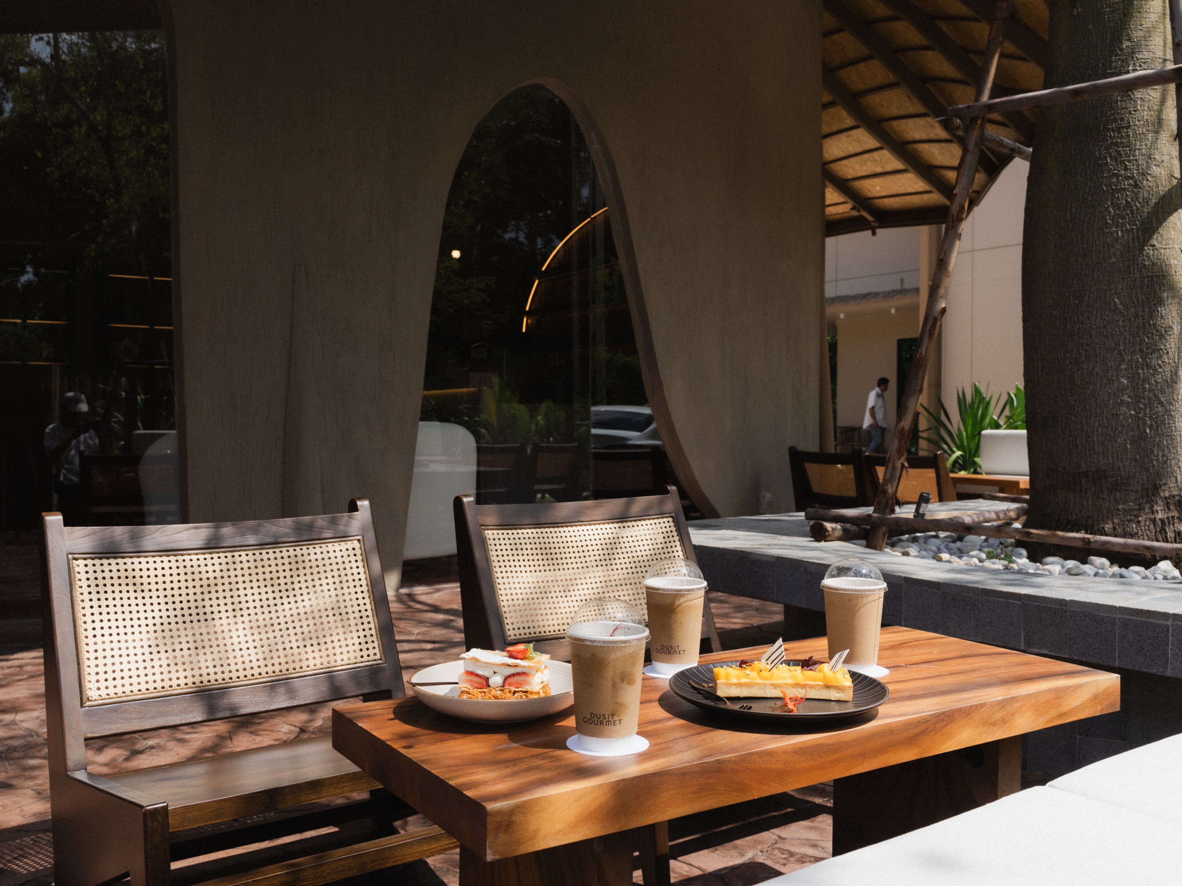 An outdoor seating area with wooden tables and chairs, two plates of food, and two drinks labeled 'DUSIT GOURMET'.