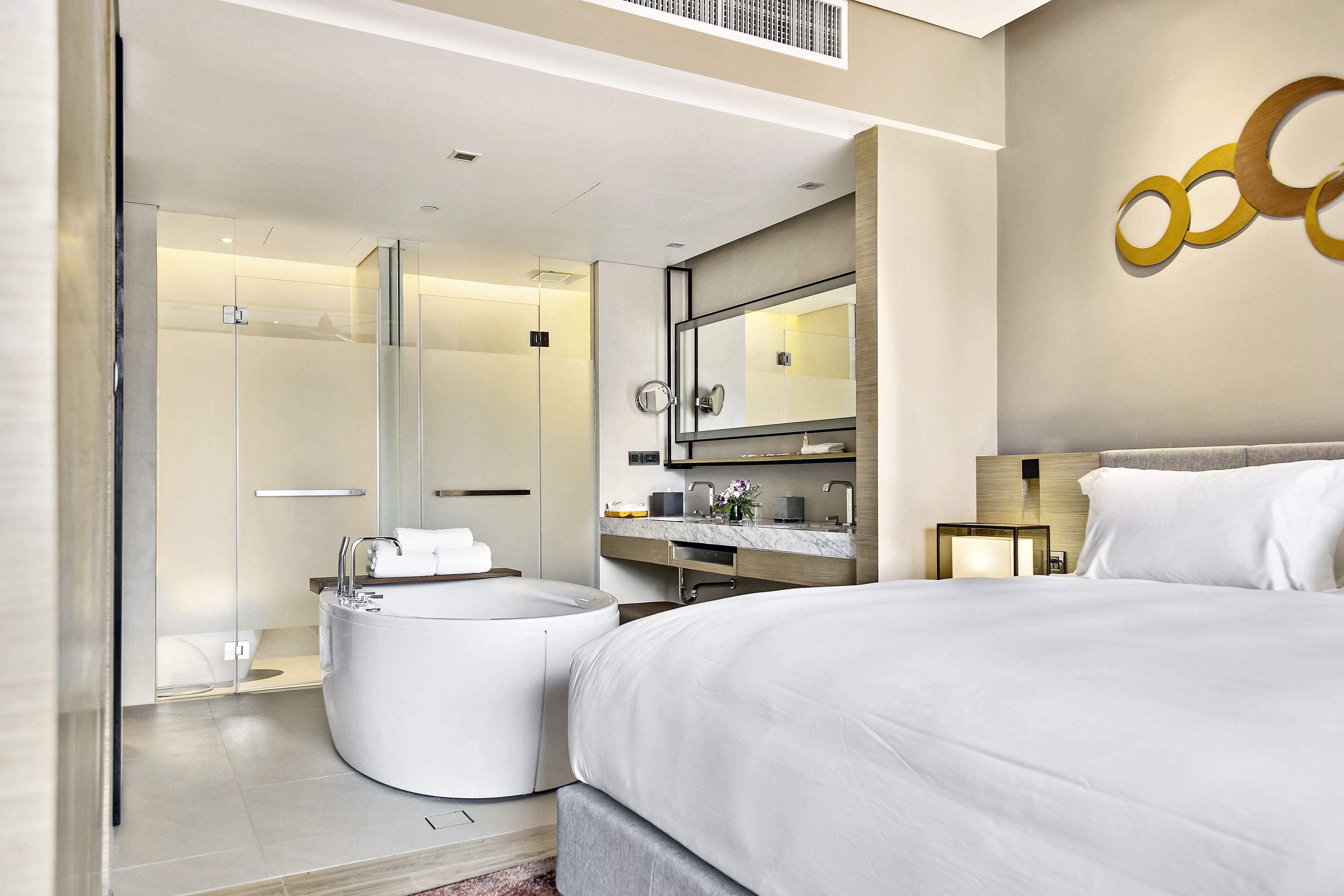 Una habitación de hotel moderna con una cama grande, una bañera redonda, un vestuario de ducha y un tocador con espejo.