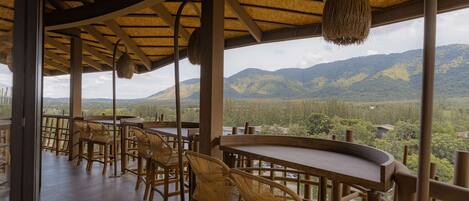 A covered outdoor seating area with wicker chairs and tables, overlooking a mountainous landscape.