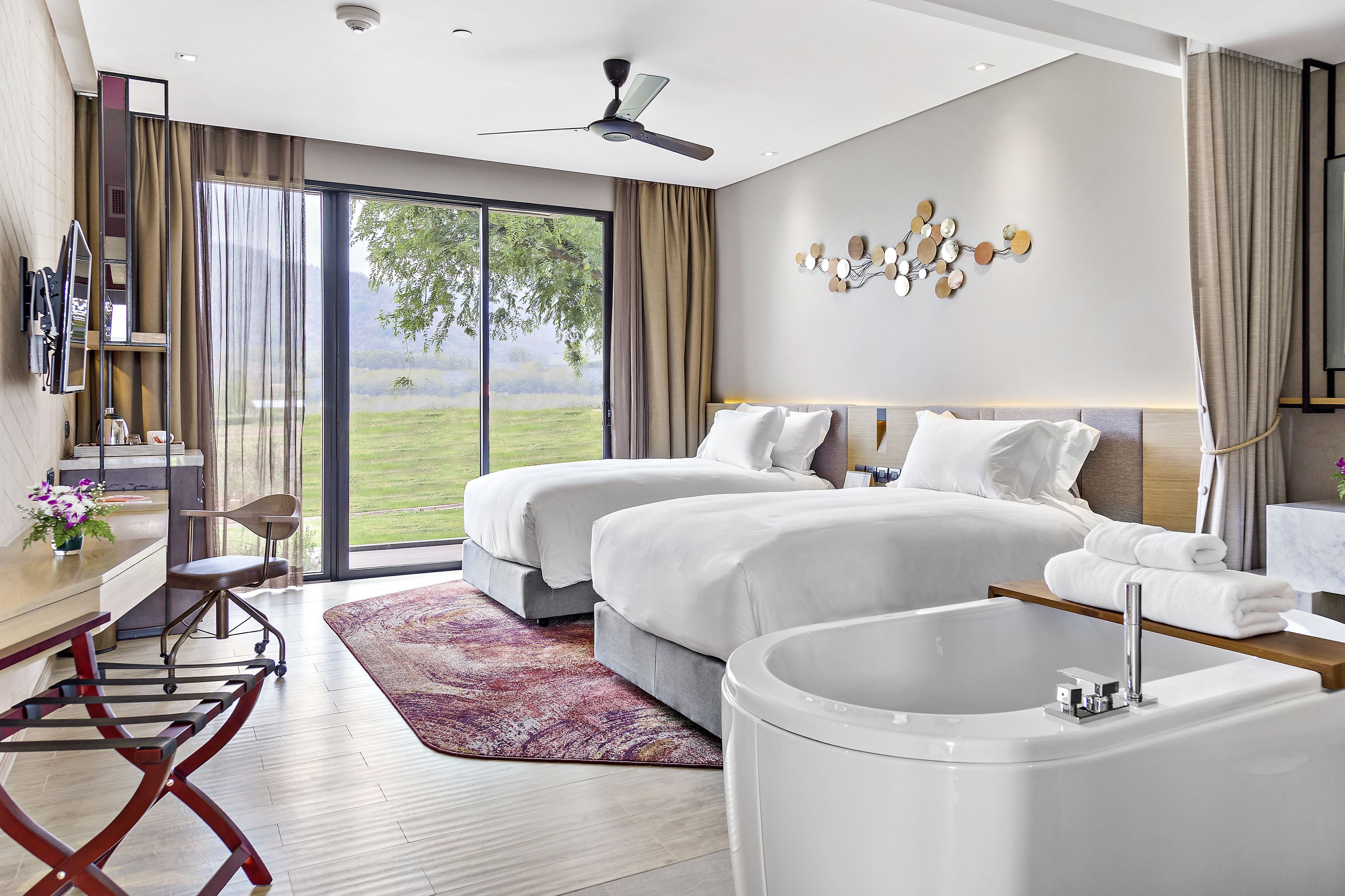 A modern hotel room with two beds, a large window with a view of a green landscape, a ceiling fan, and a bathtub.