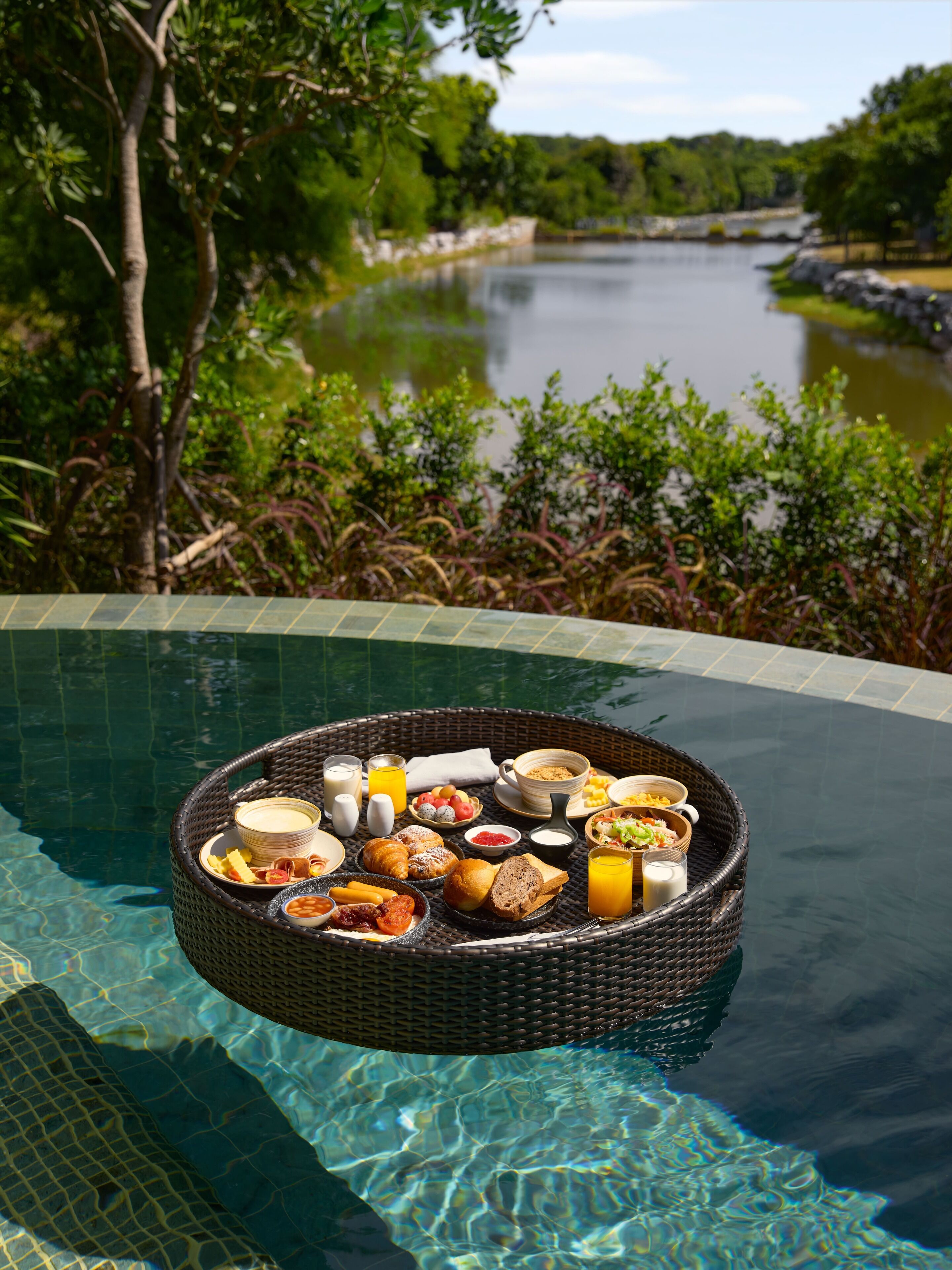 Una cesta de mimbre llena de productos para el desayuno flotando en el borde de una piscina, rodeada de exuberante follaje y un cuerpo de agua tranquilo.