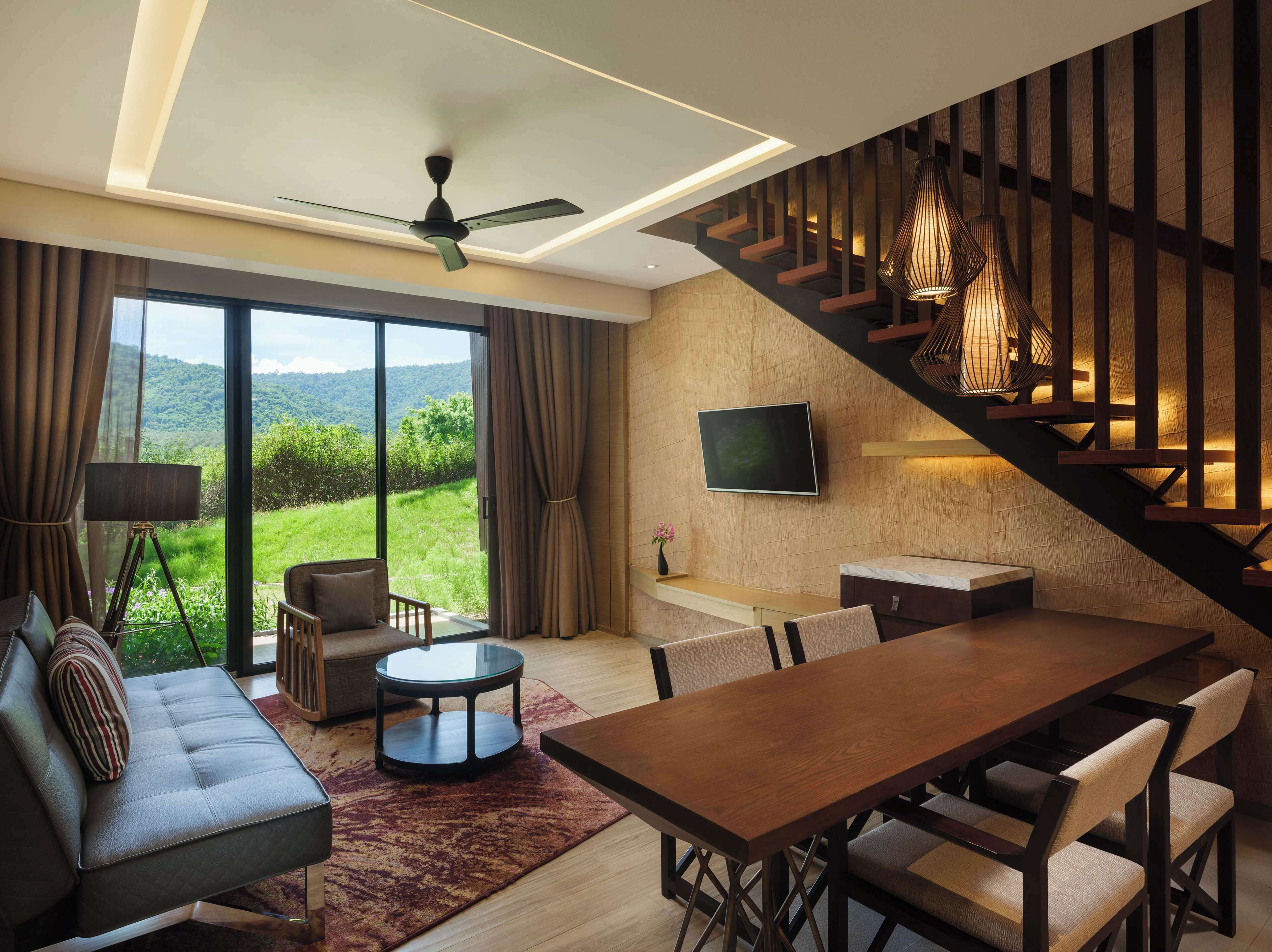 A modern living room with a sofa, a dining table, and chairs. There is a TV mounted on the wall, a ceiling fan, and a staircase with wooden railings.