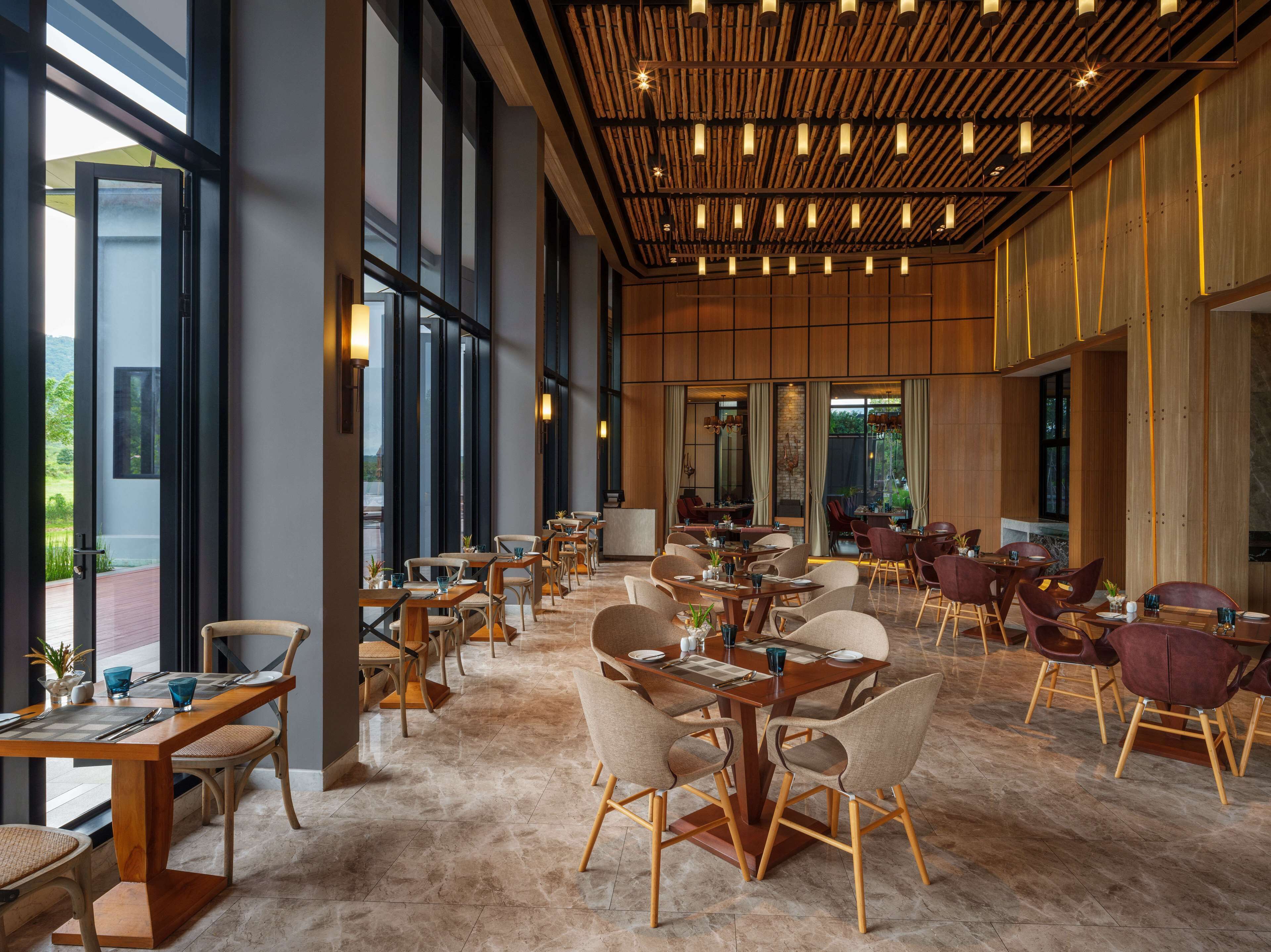 A spacious dining area with multiple tables and chairs, large windows, and a wooden ceiling with hanging lights.