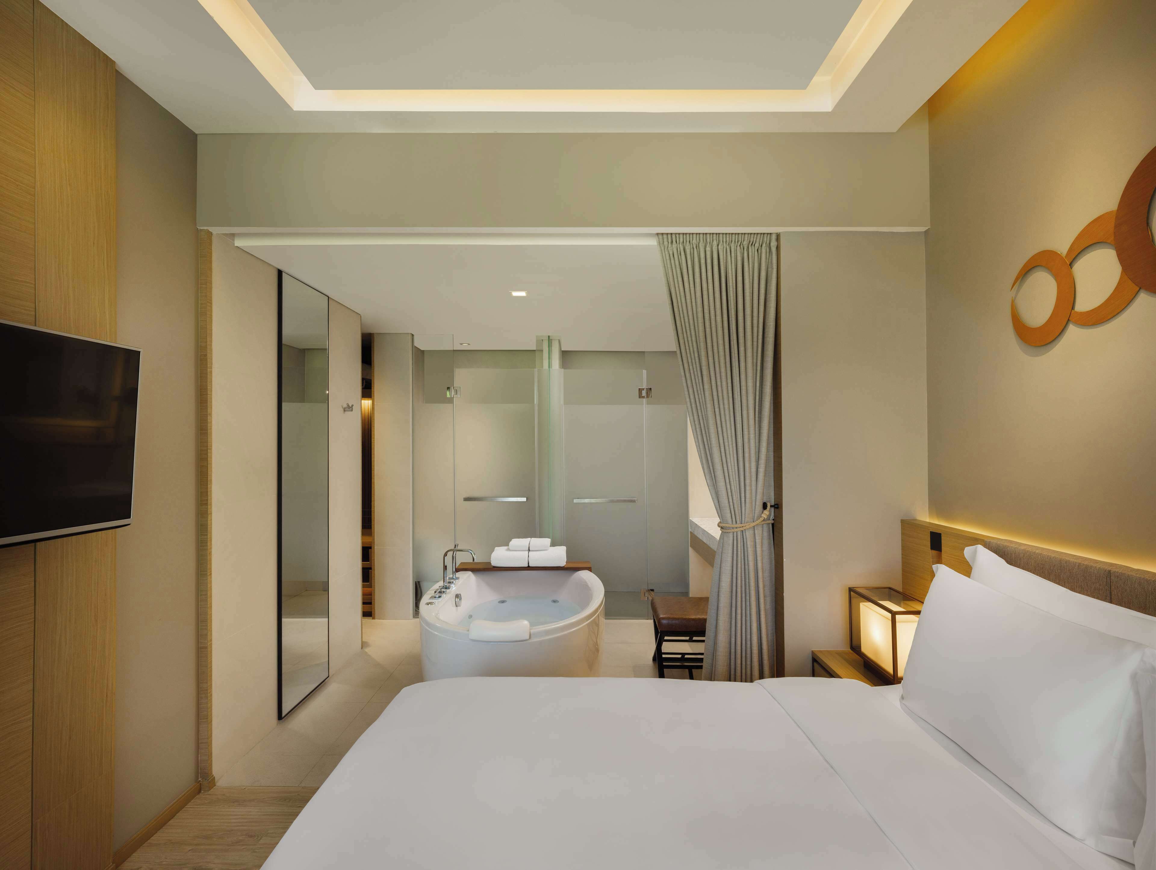 A modern hotel room with a large bed, a built-in bathtub, a TV, and a mirror.