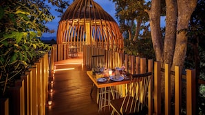 An outdoor dining area with a wooden deck, a thatched-roof gazebo, and a view of the mountains in the background. - dusitD2 Khao Yai (Pak Chong)
