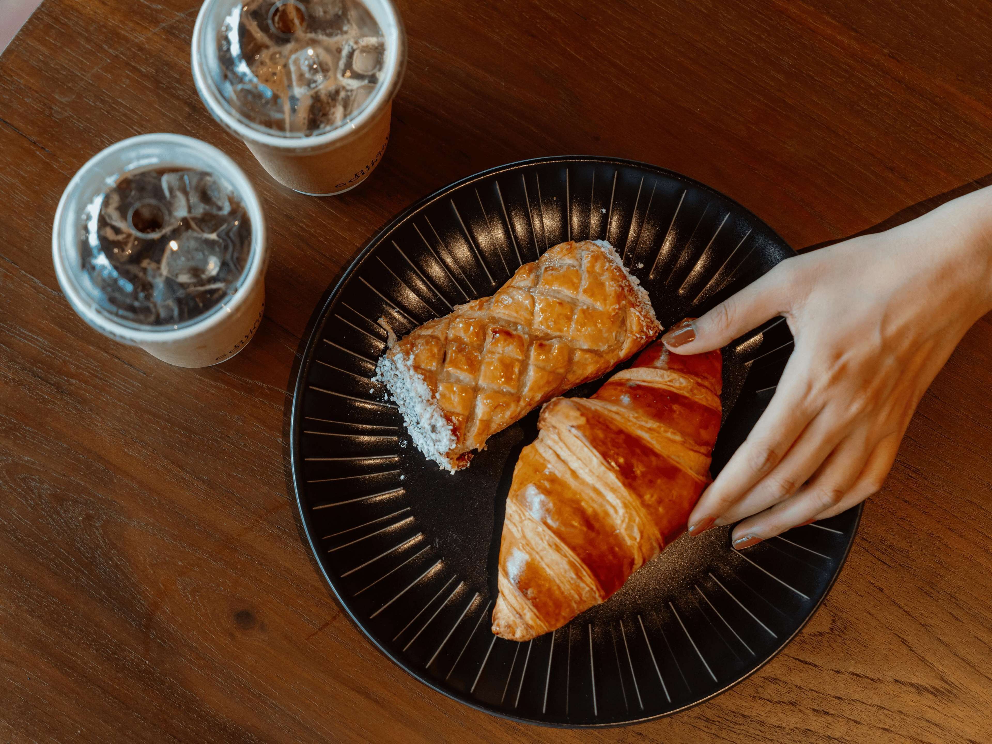 Un plato negro con dos croissants y una mano alcanzando uno. Hay dos vasos con cubitos de hielo sobre una mesa de madera.